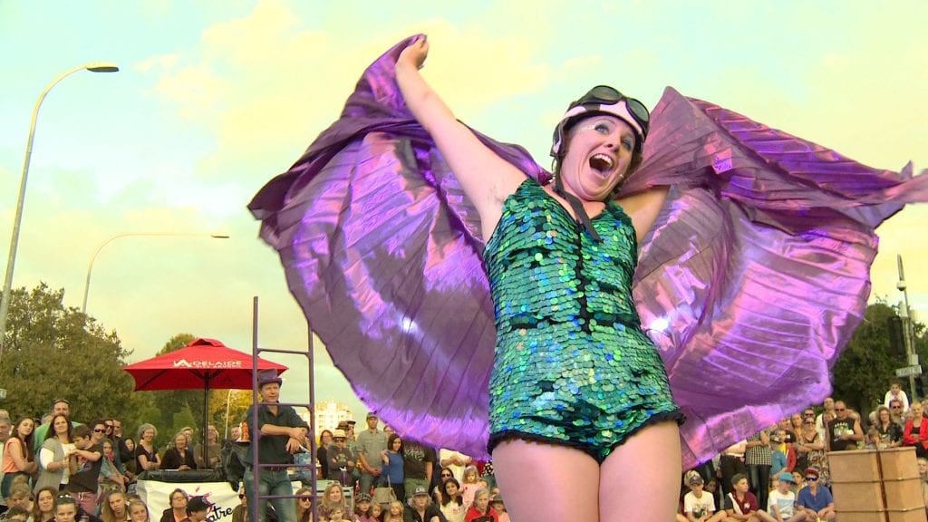 A joyful performer in a shimmering green sequin outfit and purple wings spreads her arms wide on an outdoor stage. She wears a helmet and goggles, smiling brightly as a crowd watches in the background under a golden sky.