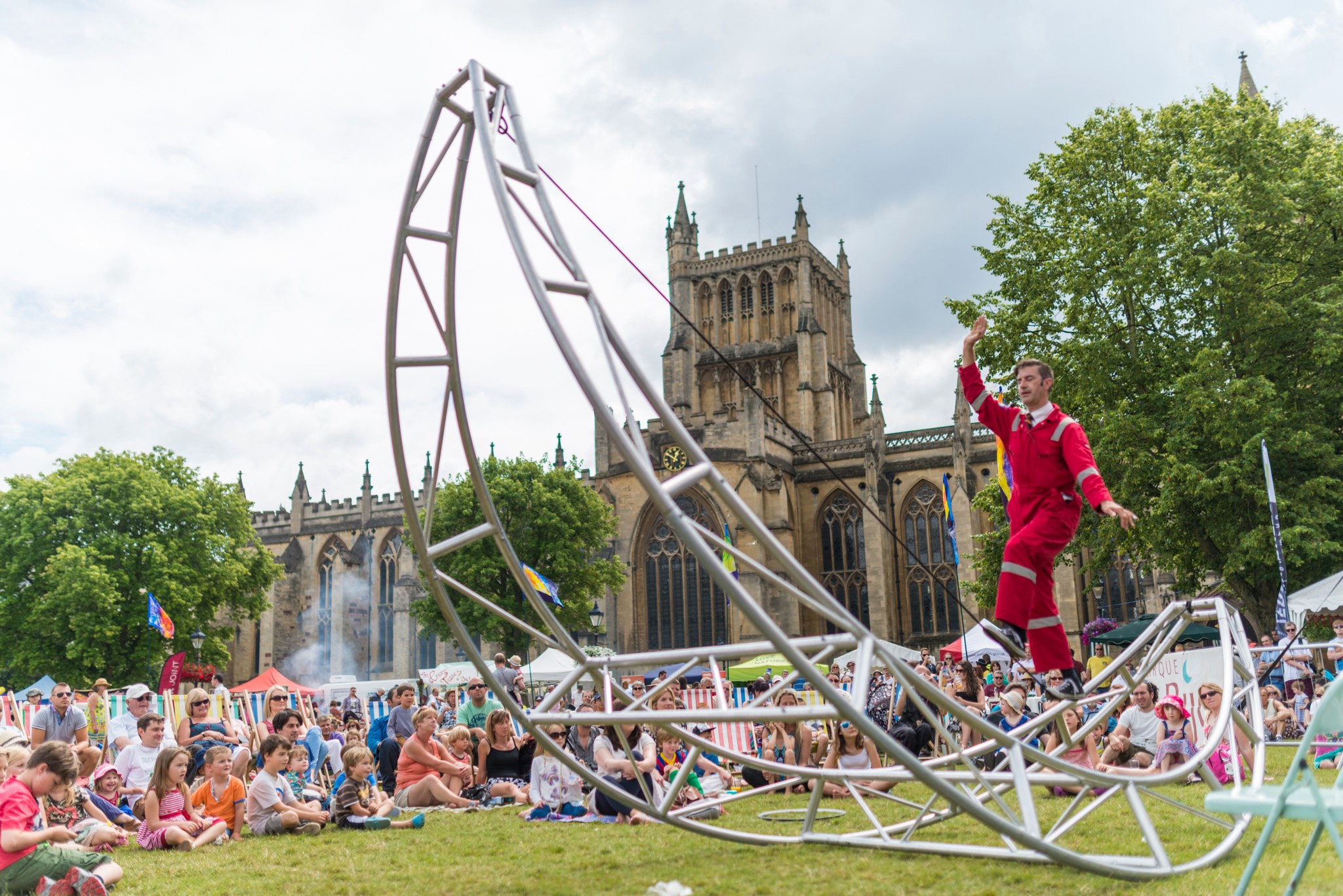 A male performer in a red jumpsuit balances on a curved metal structure during an outdoor show at a festival. A large audience, including families with children, watches from the grass. In the background, a historic cathedral rises above the crowd, adding dramatic contrast to the performance scene.