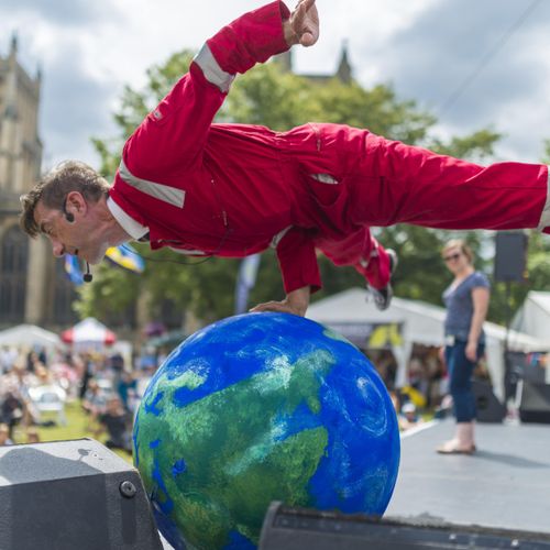 A street performer in a red jumpsuit balances on one hand atop a large painted globe, mid-act, with a historic cathedral and a festival crowd visible in the background under a partly cloudy sky.