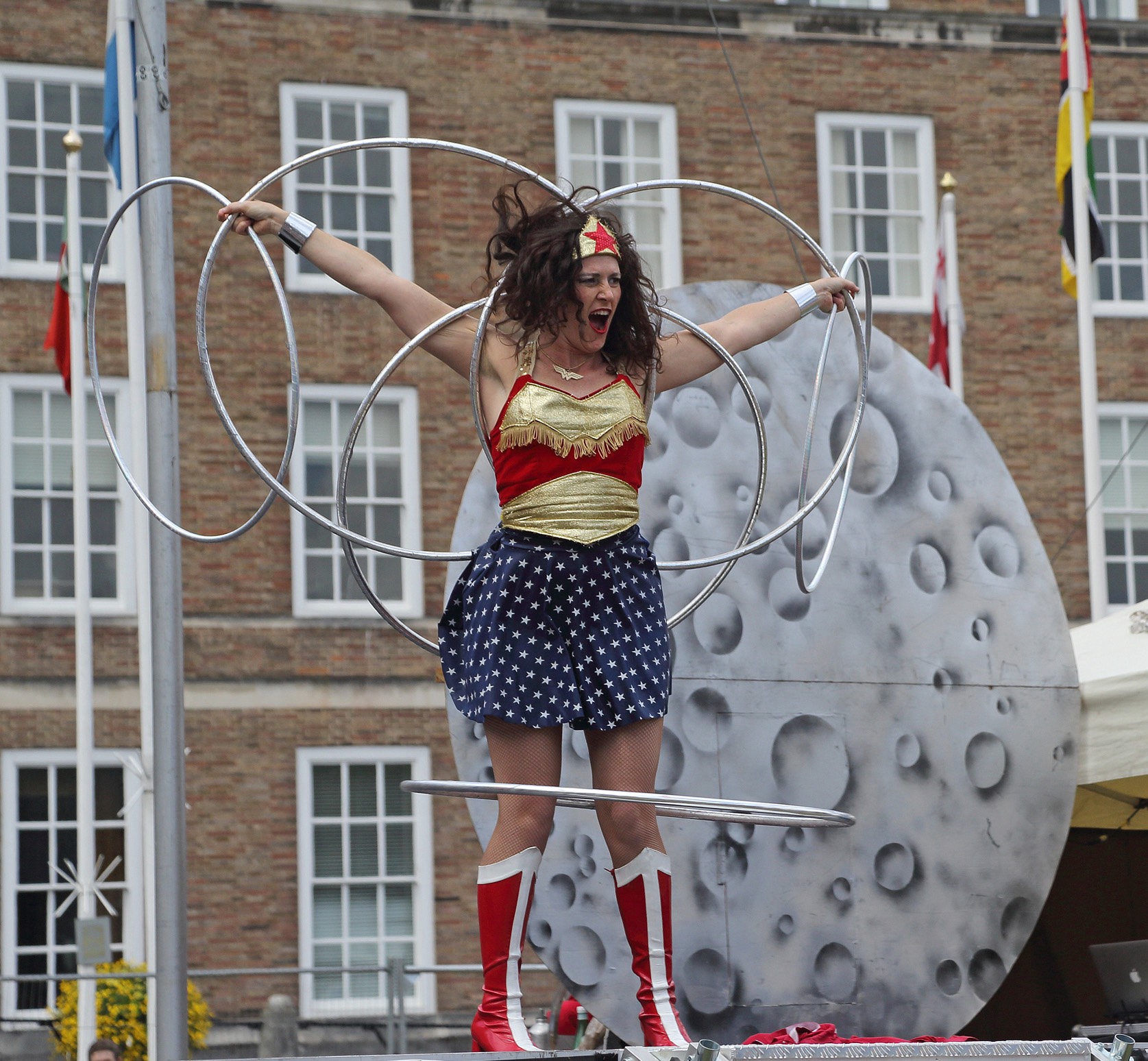 A female performer dressed as Wonder Woman hula-hoops with more than a dozen red and silver hoops on an outdoor stage. She wears a gold top, blue star-patterned skirt, and red-and-white boots, smiling confidently with arms raised. Behind her is a large red rocket prop and a crowd of spectators in front of a historic brick building.