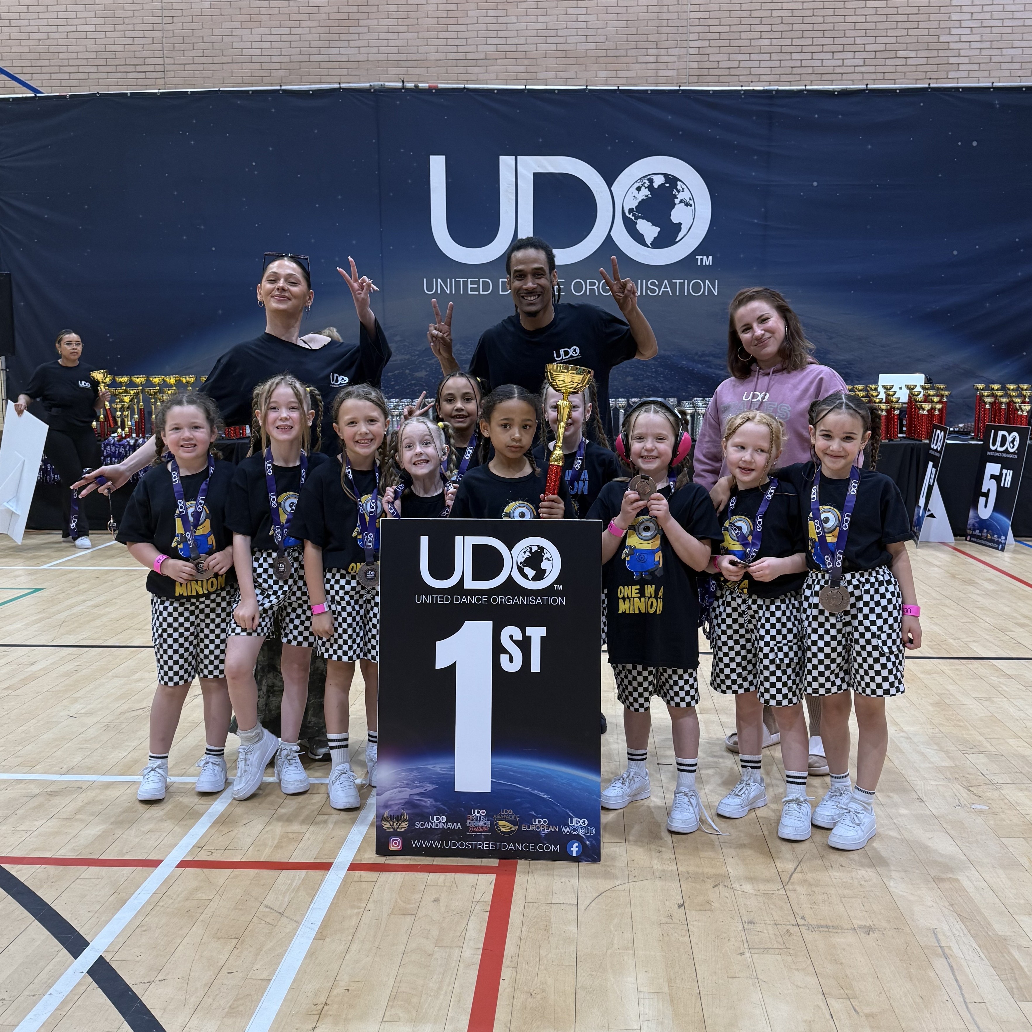 A group of young dancers celebrate winning first place at a United Dance Organisation (UDO) competition. They pose proudly with their trophy, medals, and a large sign reading "1st" in a sports hall, flanked by their smiling coaches. All wear matching black-and-white checkered outfits and "One in a Minion" T-shirts.