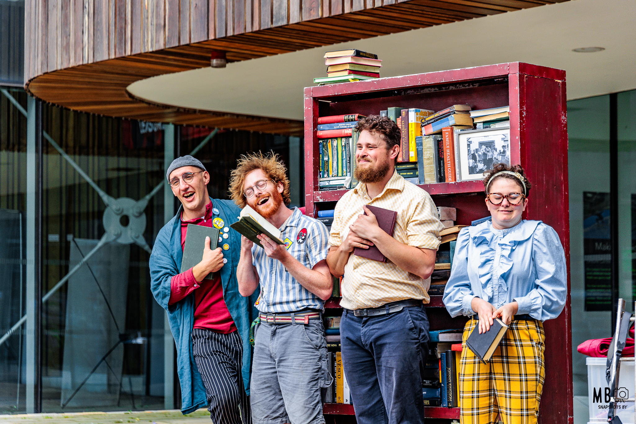 Four performers stand in front of a large red bookcase filled with books, each holding a book and expressing animated, theatrical emotions. They are dressed in quirky, colorful clothing, suggesting a humorous or whimsical outdoor performance. The setting appears to be outside a modern building, with glass and wood architectural elements in the background.