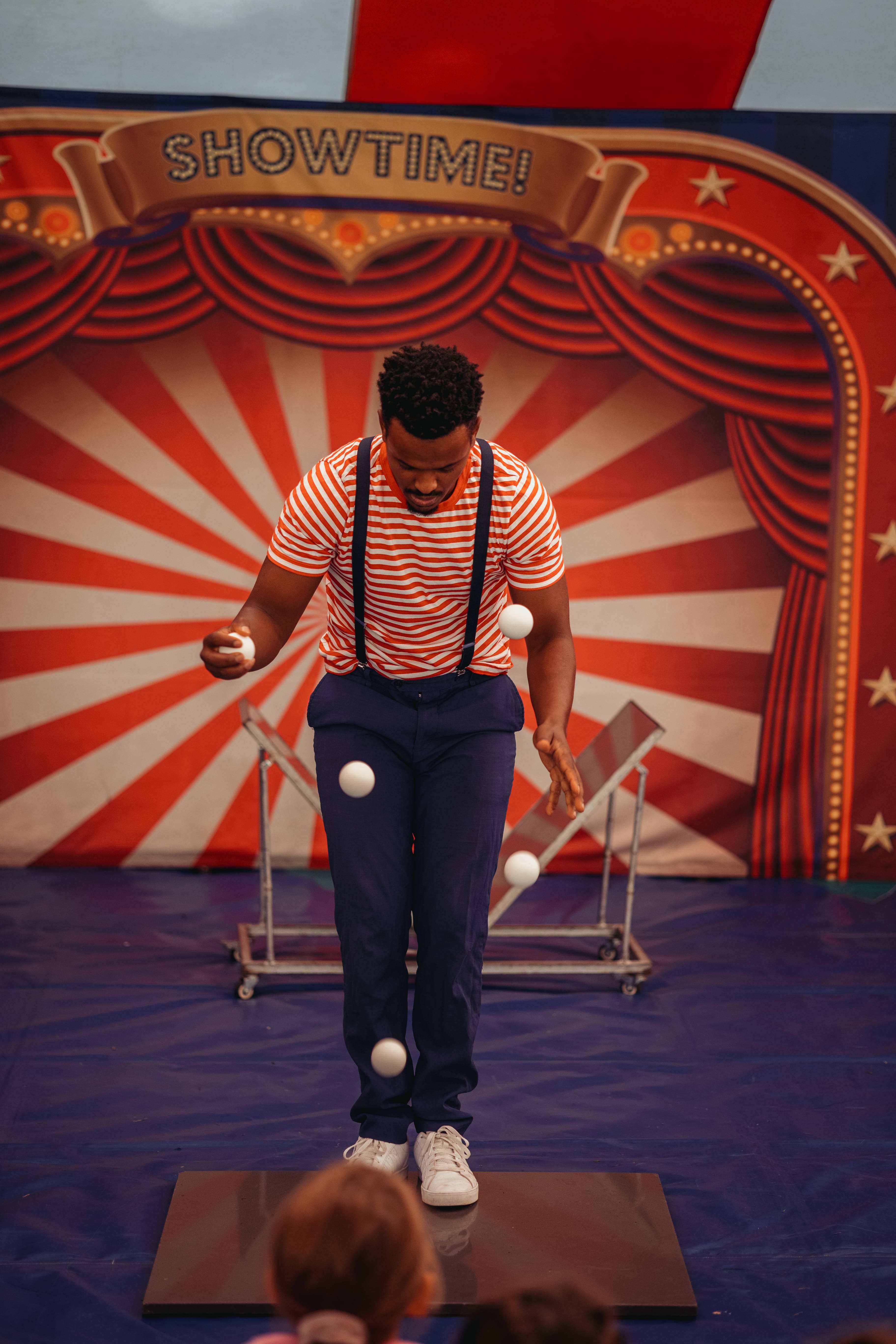 A circus performer in a red and white striped shirt, navy trousers with suspenders, and white trainers is juggling five white balls in front of a vibrant "Showtime!" backdrop featuring red and white rays and decorative curtains. A small audience, including a child with a ponytail, watches the performance on a blue mat.