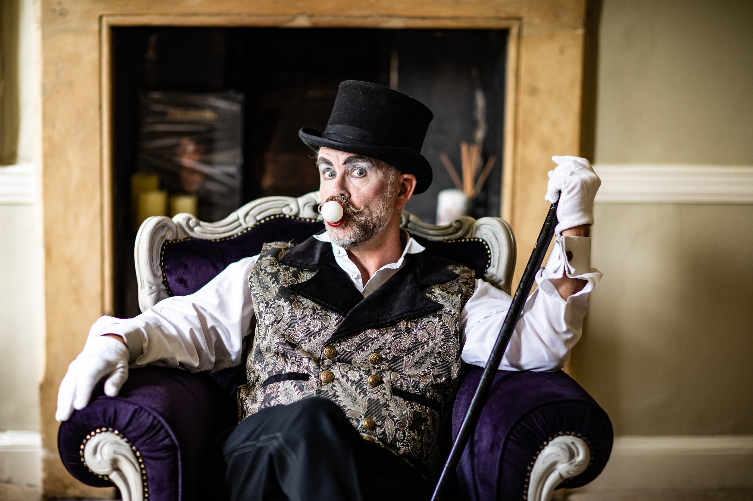 A performer dressed in vintage circus attire sits confidently in a plush purple armchair. He wears a top hat, white gloves, a patterned waistcoat, and holds a black cane. His face is painted with theatrical makeup, and a small white clown nose adds to his expressive look. The setting appears to be an elegant room with a fireplace in the background.