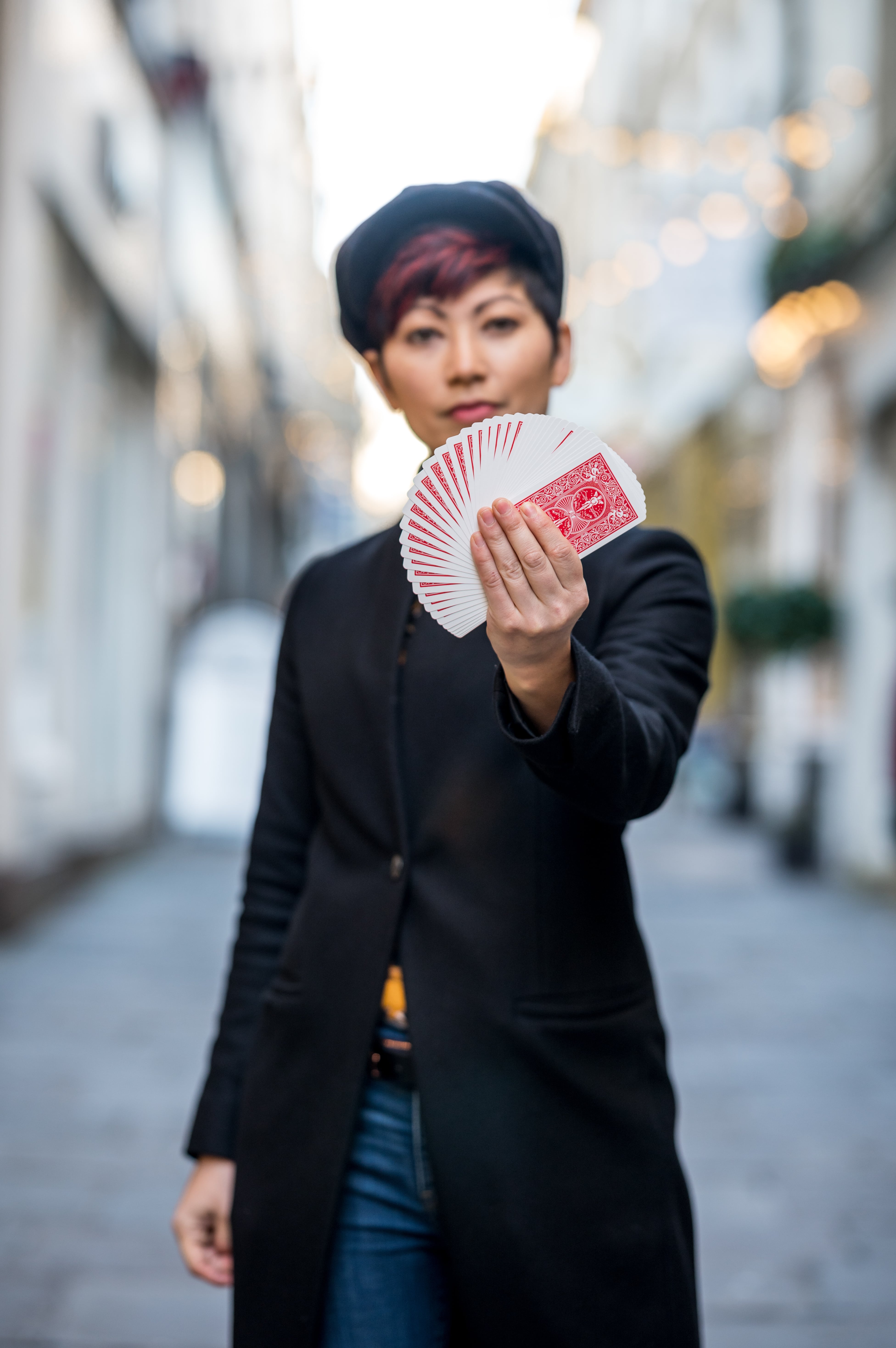 A street magician holds out a perfectly fanned deck of red playing cards towards the camera. They wear a black coat, blue jeans, and a black flat cap, standing confidently in a blurred urban alley with soft lights in the background.