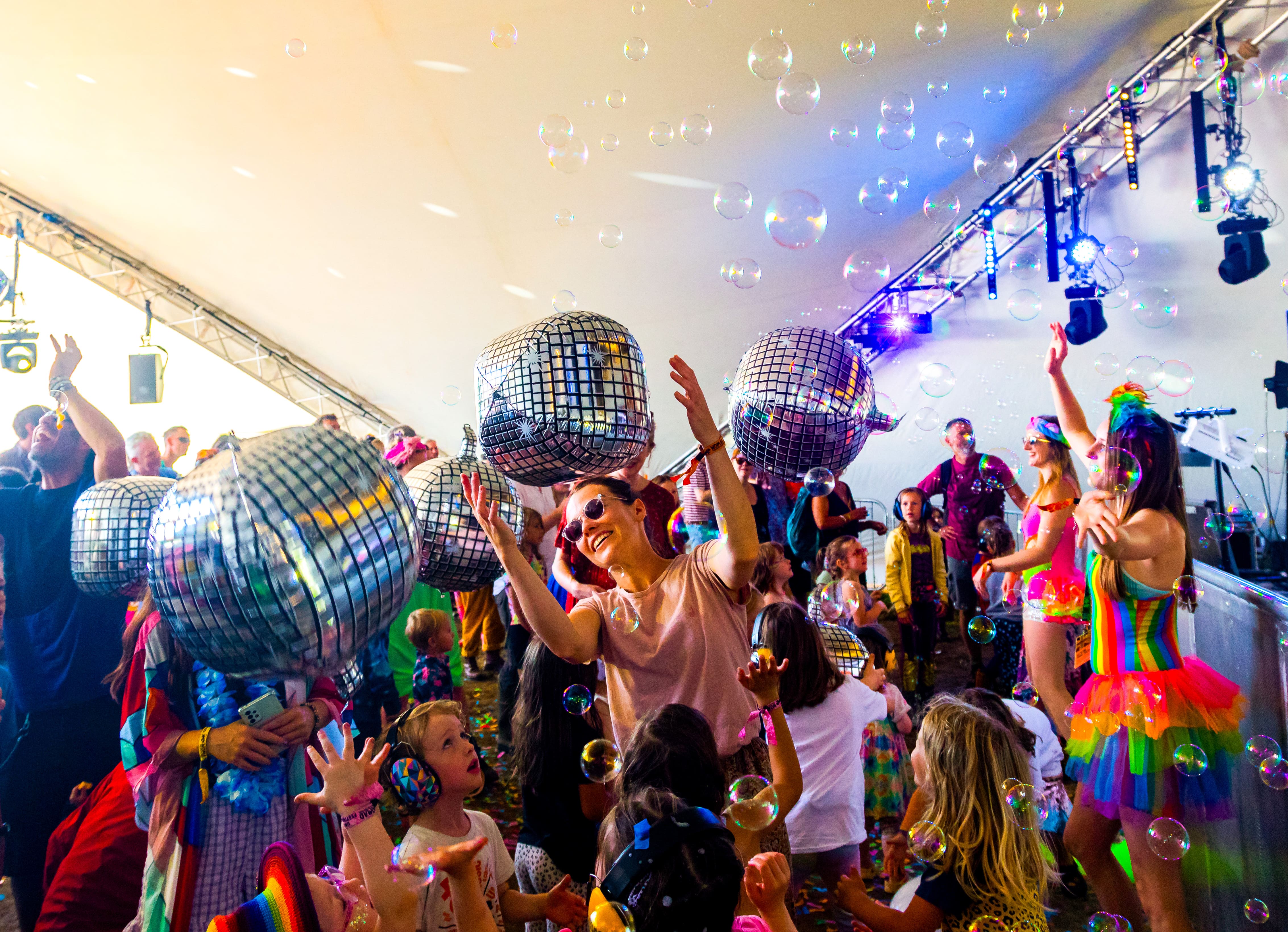 A lively festival scene under a big tent filled with bubbles, inflatable disco balls, and people dancing. Adults and children in colourful outfits, including rainbow costumes and sunglasses, reach up joyfully as music plays and bubbles float through the air. Bright lights and stage equipment enhance the energetic atmosphere.