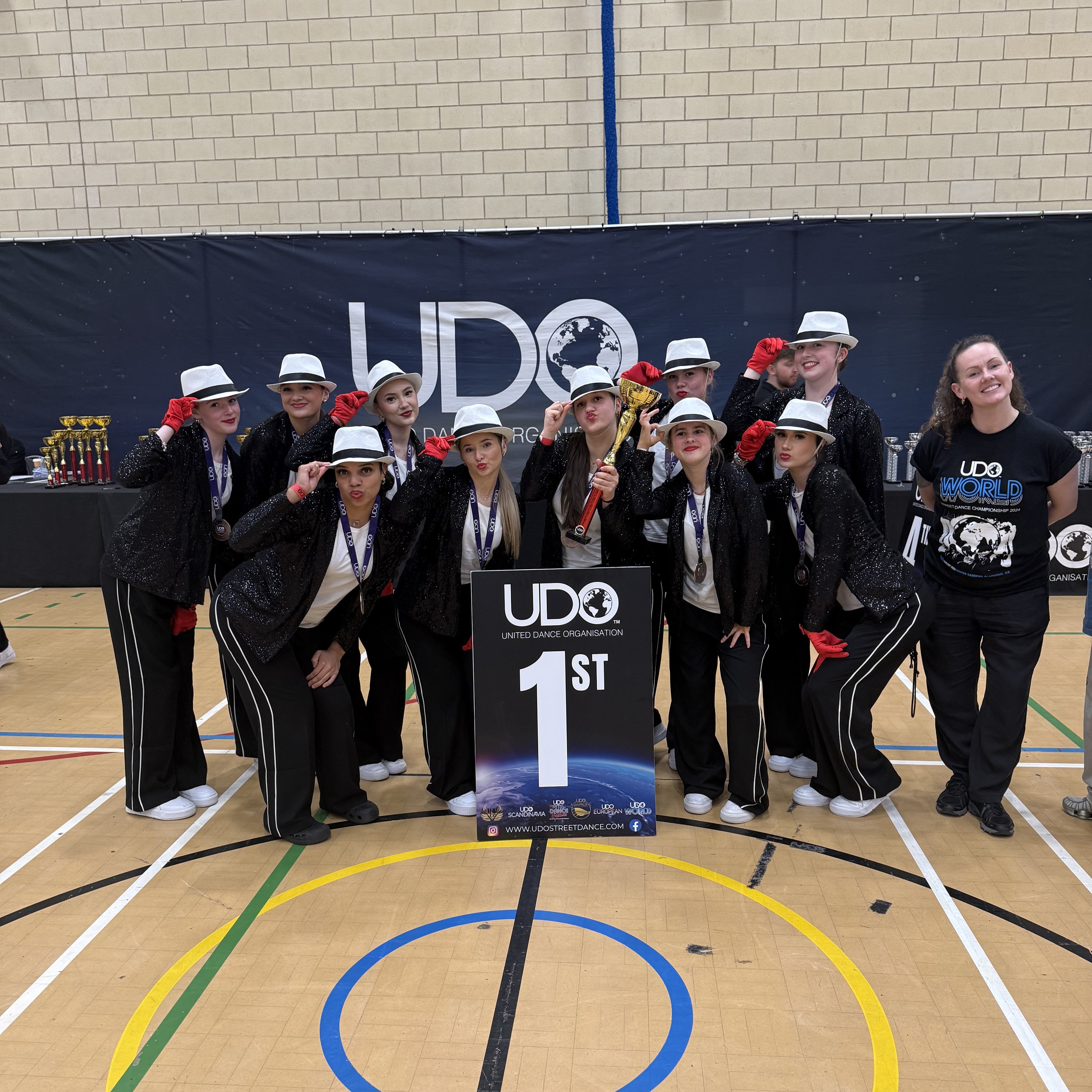 A dance team poses proudly indoors after winning first place at a UDO (United Dance Organisation) competition. The group wears black sparkly jackets, white hats, white shoes, and red gloves, standing behind a large sign that reads “UDO 1st.” A coach or team leader in a branded UDO t-shirt stands smiling at the side.