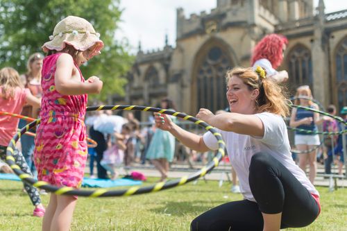 Children race down a green track as a large, cheerful crowd of families watches and cheers. The event takes place outdoors in front of a historic church building, with some people holding umbrellas, suggesting light rain. The children wear colorful outfits and joyful expressions.