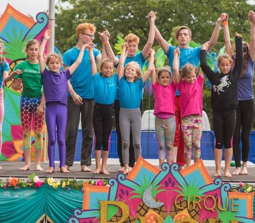 Three acrobatic performers dressed in colorful, coordinated circus-style costumes form a human pyramid at an outdoor festival. Two performers stand on the grass holding up a third who balances confidently on their shoulders with arms outstretched. A lively crowd and fairground structures are visible in the background.