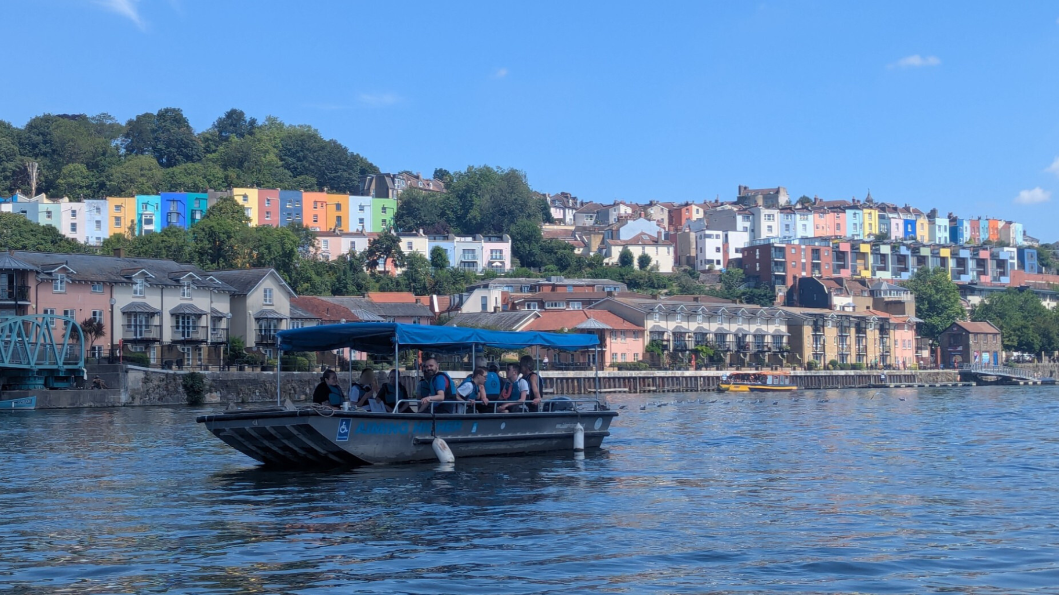 A boat carrying people in life jackets travels along Bristol’s Harbourside on a sunny day, with the city’s iconic colourful hillside houses in the background.