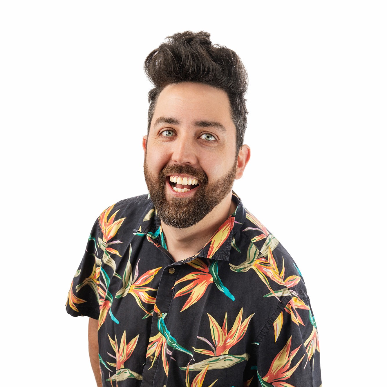 A cheerful man with a styled beard and voluminous hair smiles up at the camera. He is wearing a black shirt with a colourful tropical bird-of-paradise flower print, standing against a white background with his hands in his pockets.