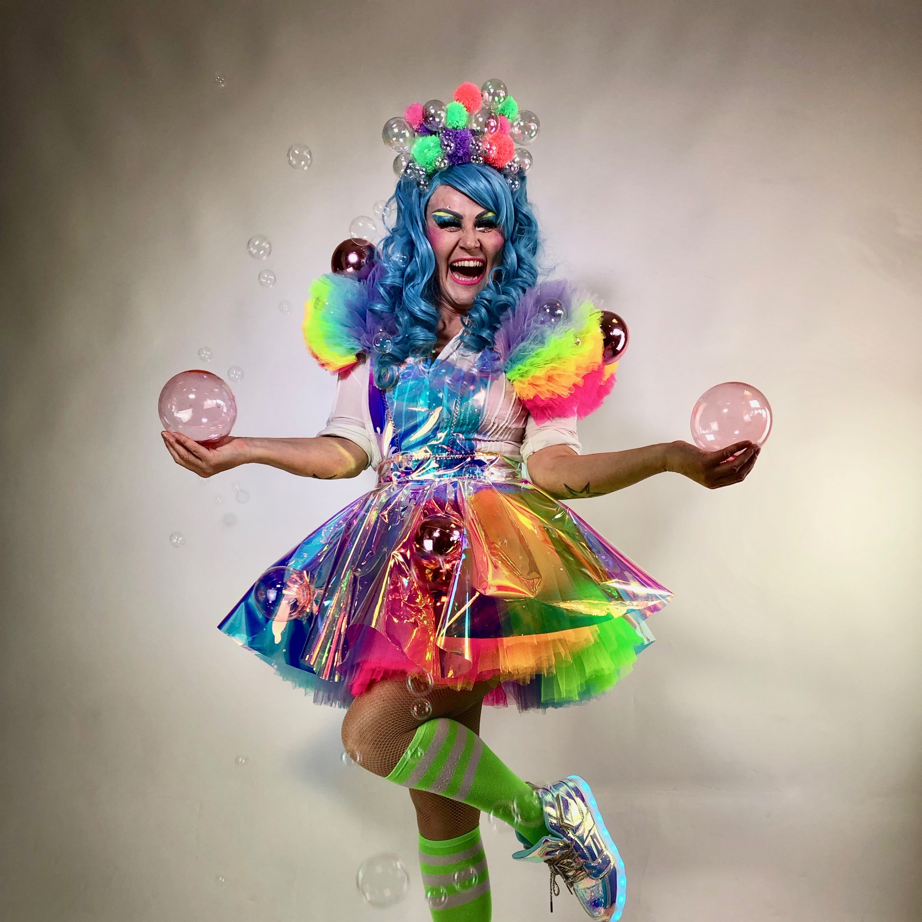 A vibrant performer with bright blue curly hair and a beaming smile poses against a plain backdrop. They wear a rainbow-colored, iridescent dress adorned with pom-poms and clear bubbles, along with neon green knee-high socks and light-up silver sneakers. They juggle pink transparent balls and are surrounded by pastel and metallic baubles on the floor, creating a whimsical, joyful scene full of color and energy.