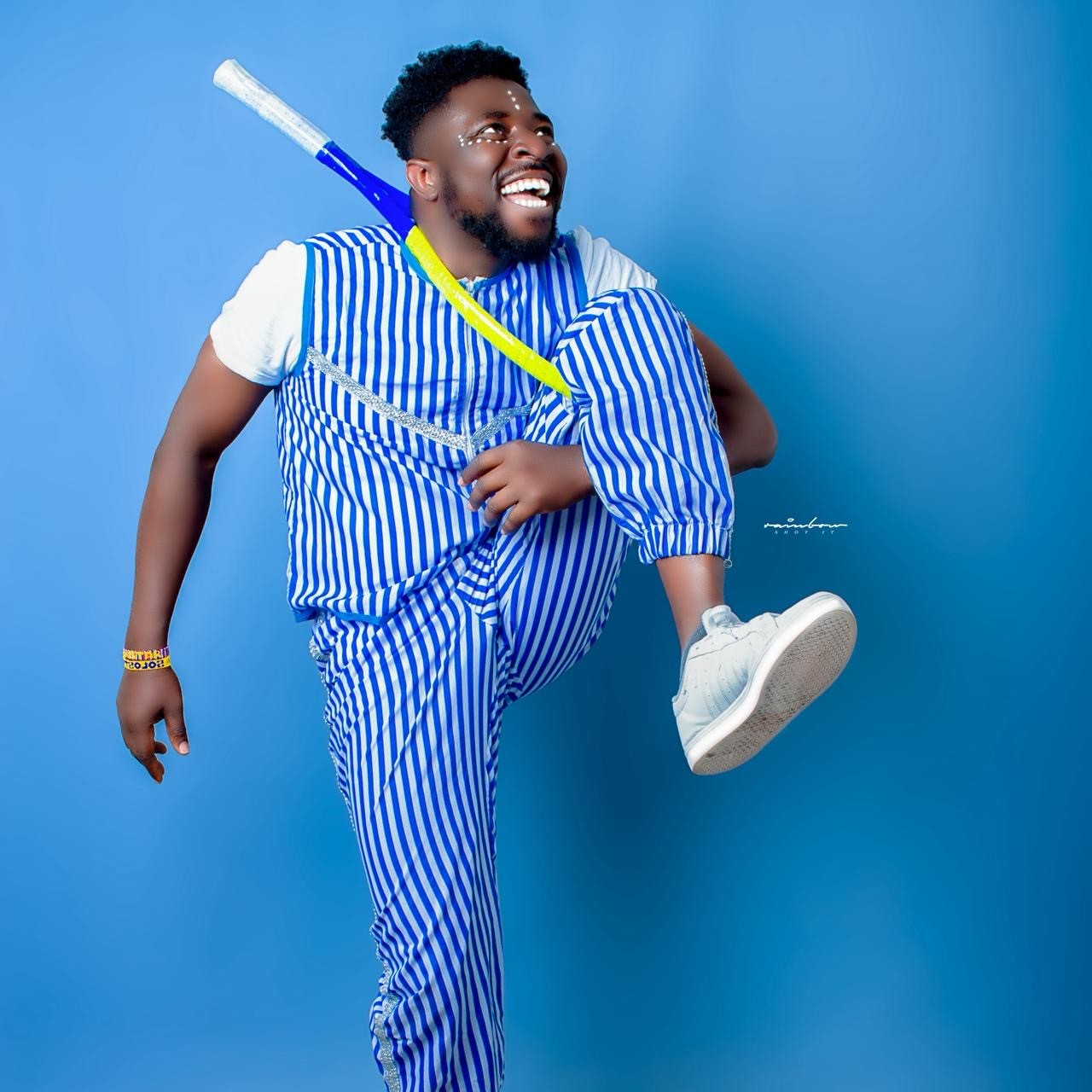 A joyful performer poses mid-motion in front of a vibrant blue backdrop, dressed in a blue and white striped outfit with matching joggers and a short-sleeved top. He wears white sneakers and carries a colorfully painted baseball bat slung over his shoulder. White face paint dots accent his cheeks, and he beams with energy and charisma.