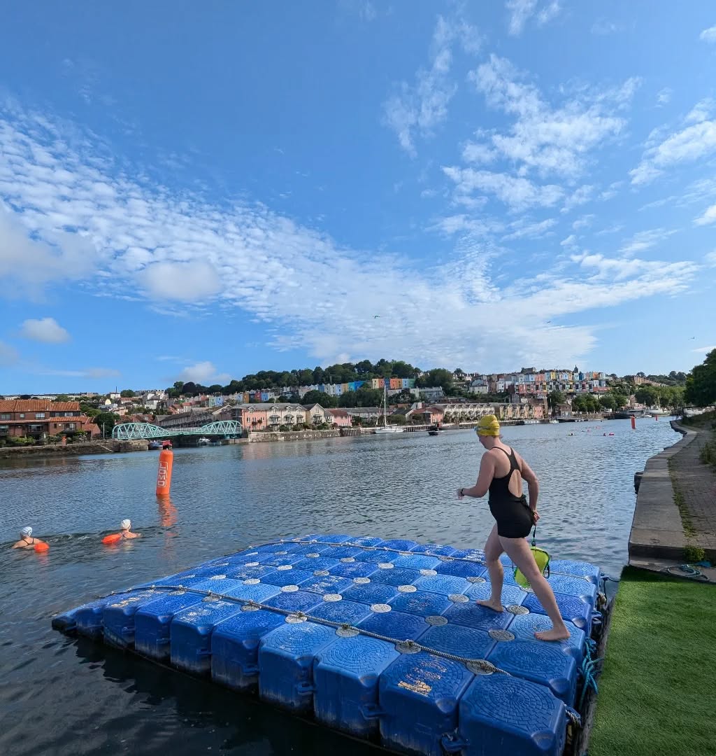 A swimmer in a black swimsuit and yellow cap steps onto a floating blue pontoon on Bristol Harbour. Other swimmers with orange tow floats are in the water, and colourful houses on the hillside form a scenic backdrop under a bright blue sky dotted with clouds.