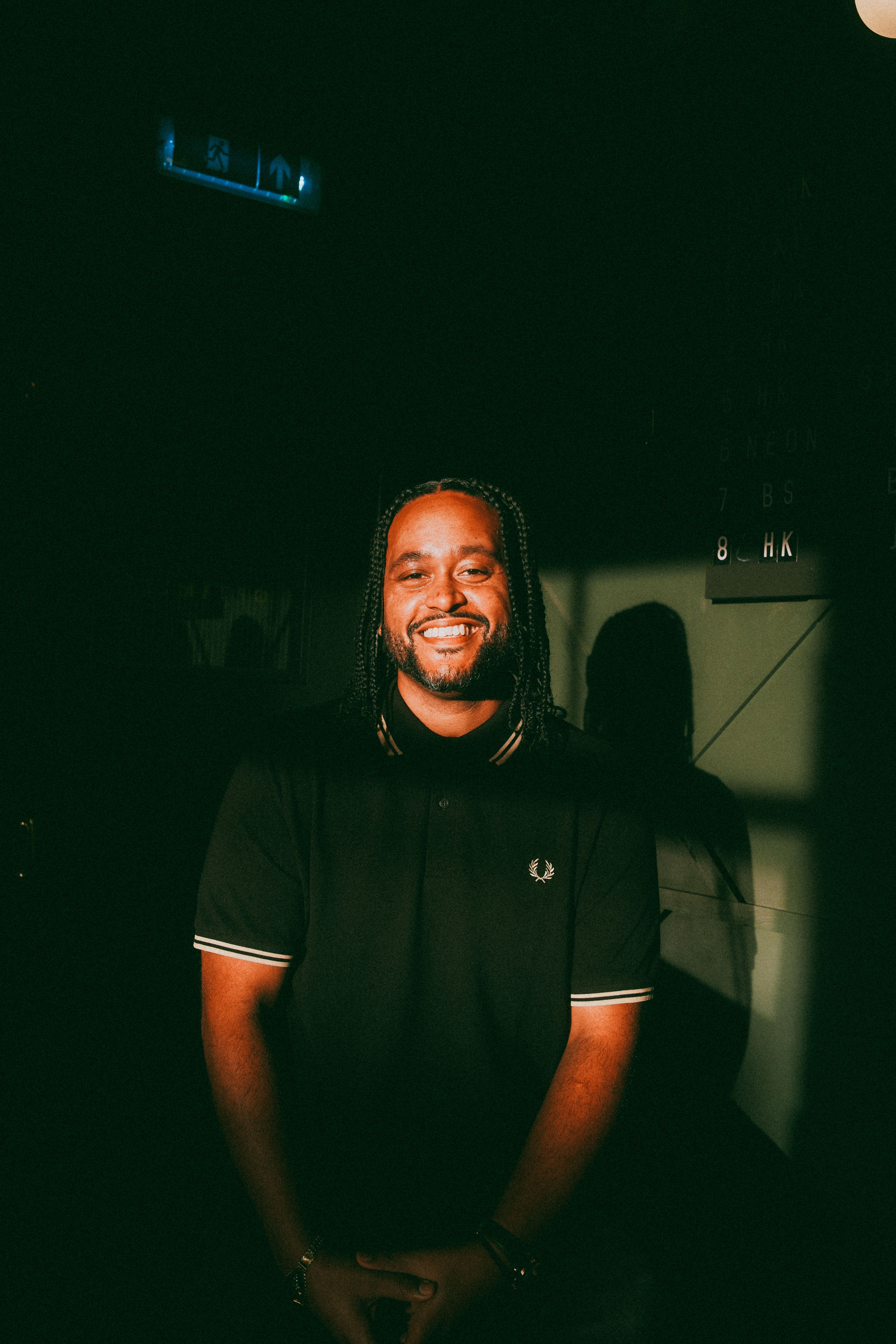 A man with braided hair smiles warmly under dramatic lighting, wearing a black Fred Perry polo shirt. A shadow is cast behind him on a dark background.