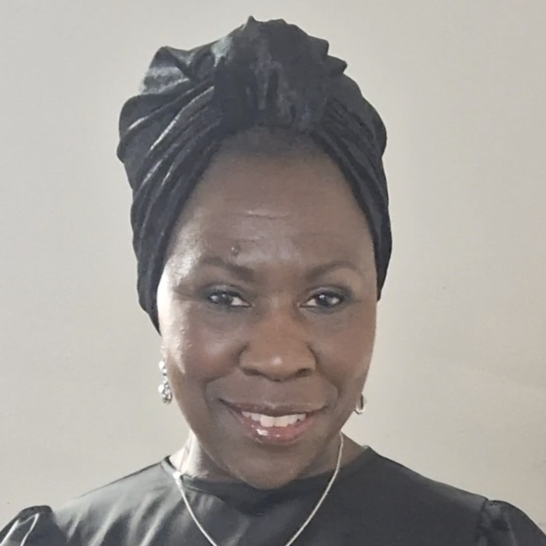 This image features a smiling woman in a satin-textured black dress and matching headwrap. She wears a delicate necklace and earrings, standing confidently against a soft neutral background.