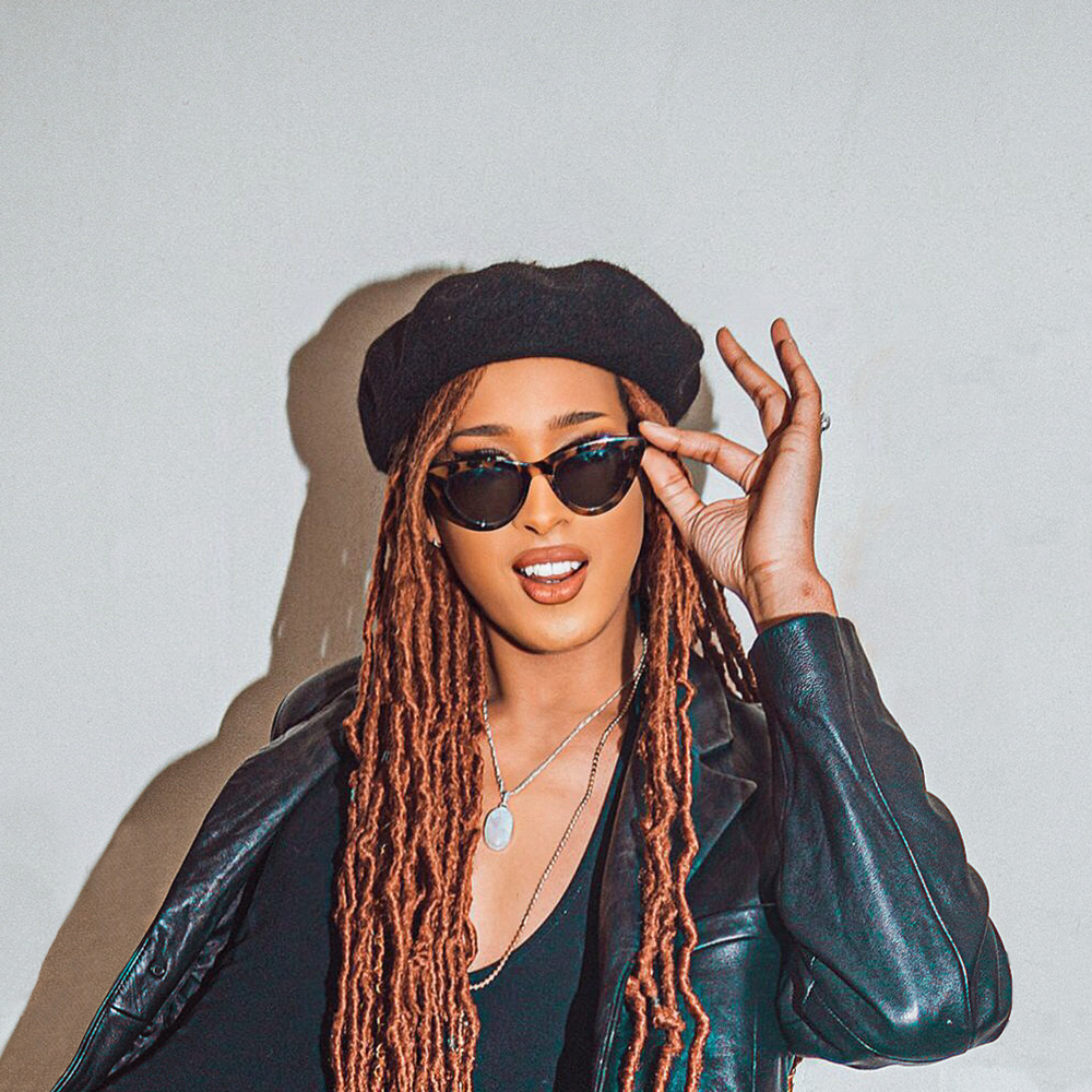 Portrait of a person wearing a black beret, sunglasses and a leather jacket, adjusting their glasses against a plain background.