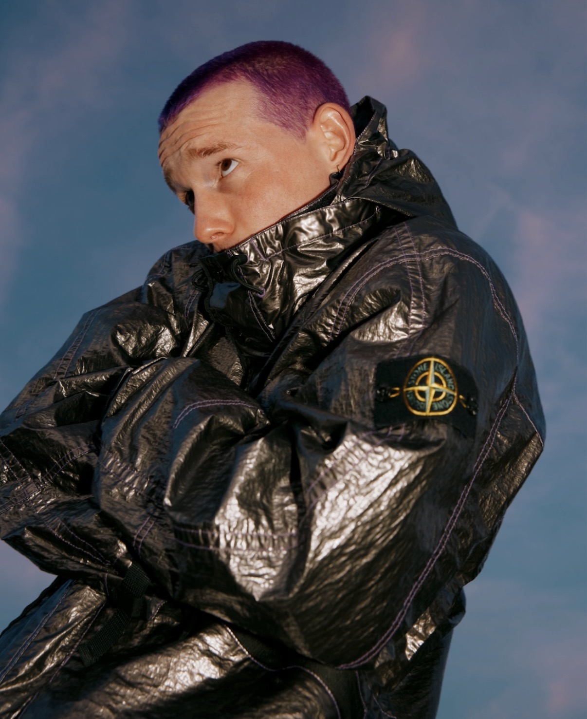 Portrait of a person with short purple hair wearing a shiny black jacket, looking to the side against a blue sky background.