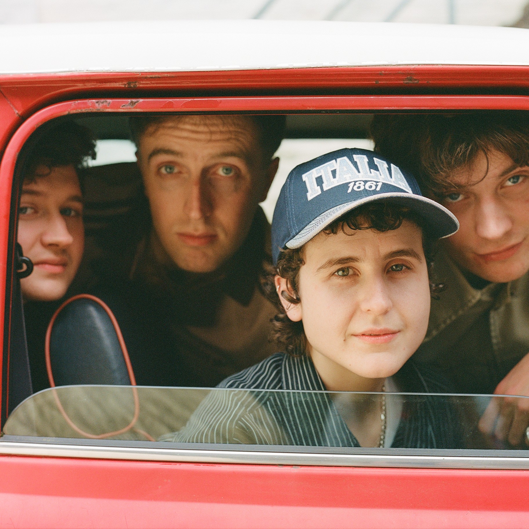 Four young people sit inside a red car, looking out through the open window, with one wearing a blue cap.