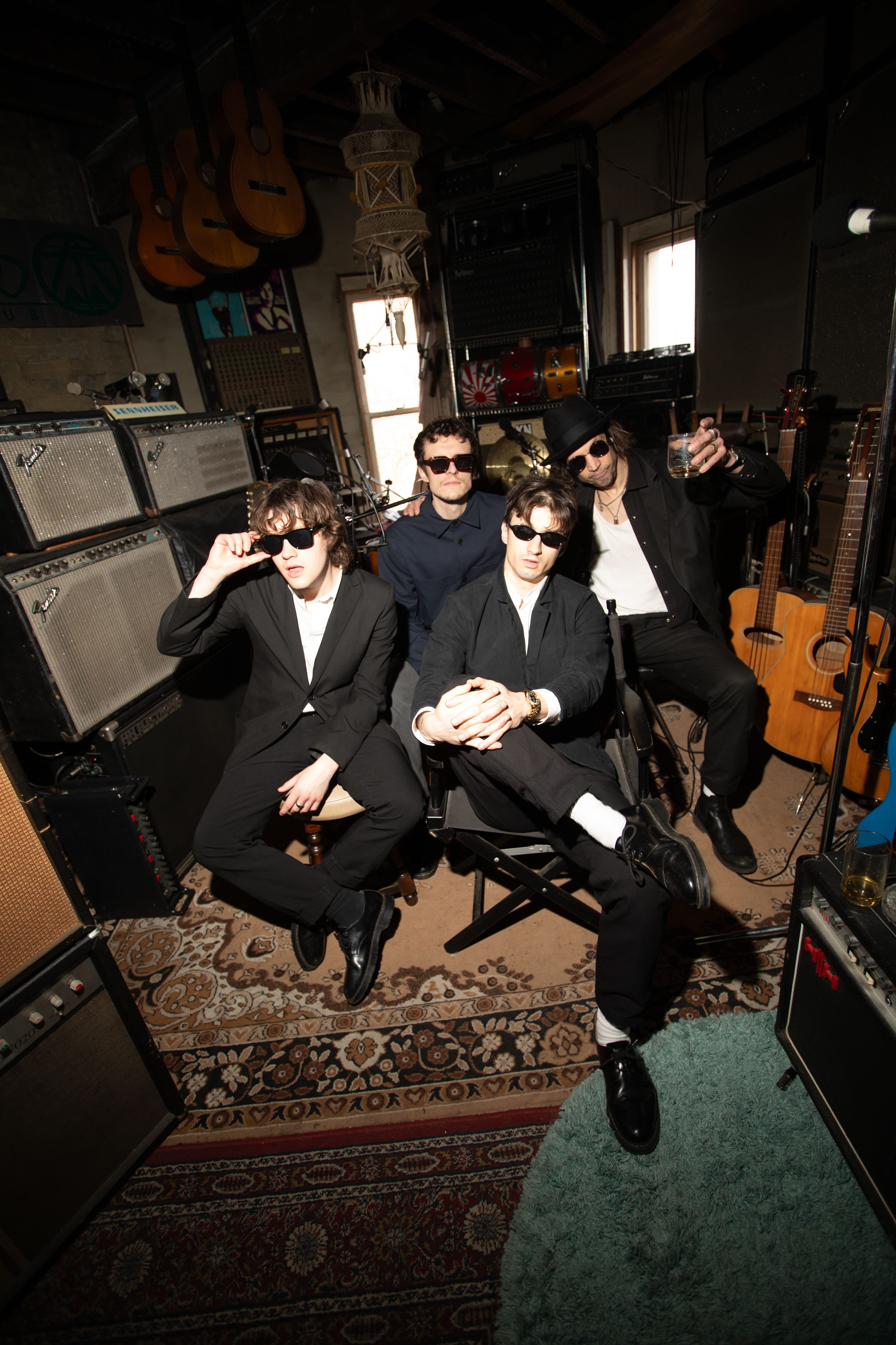 Four musicians wearing dark outfits and sunglasses sit together in a studio filled with guitars and amplifiers, posing casually.