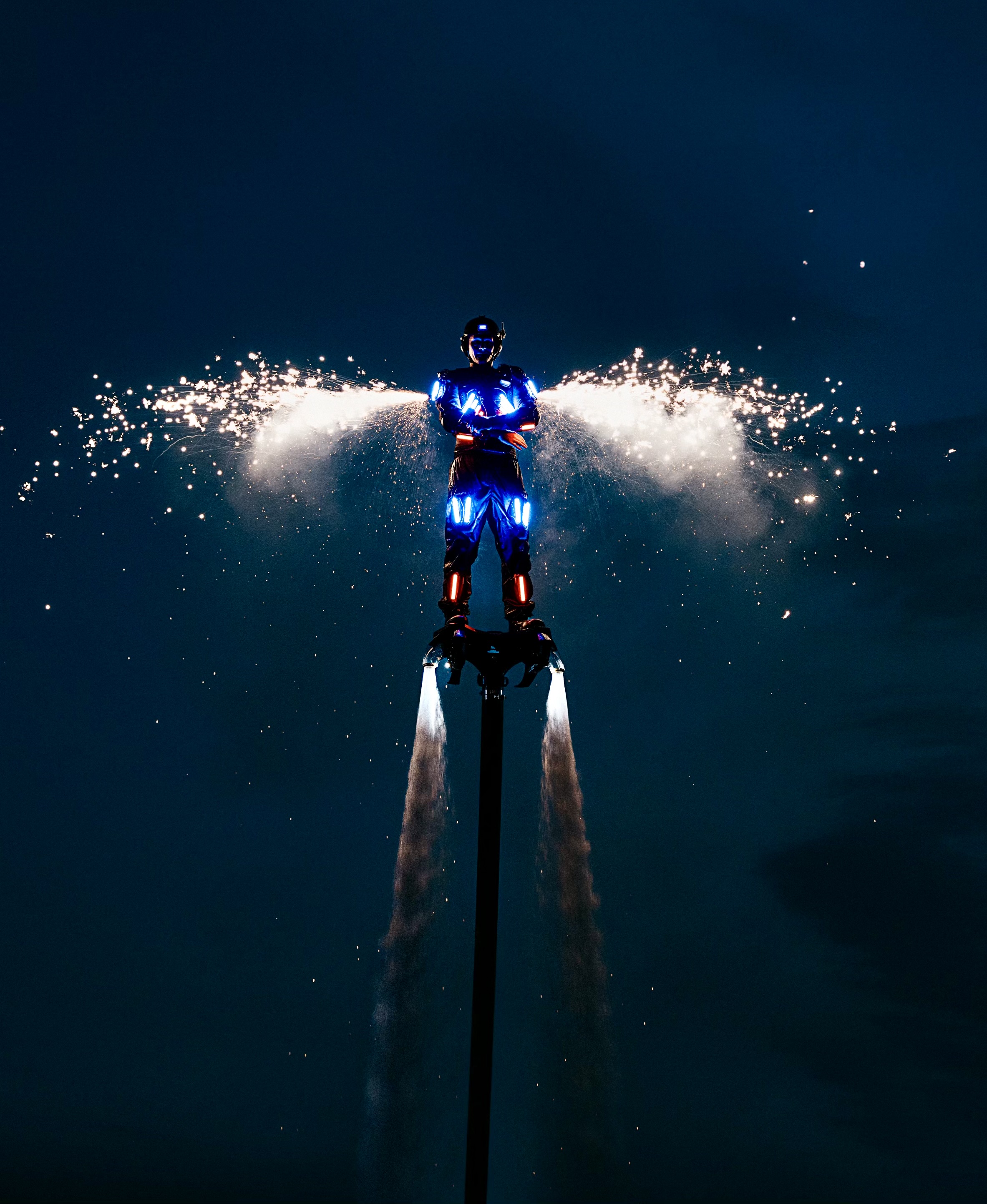 A performer in a glowing LED suit flies above water on a jet-powered device, with sparks shooting out like wings against a dark night sky.