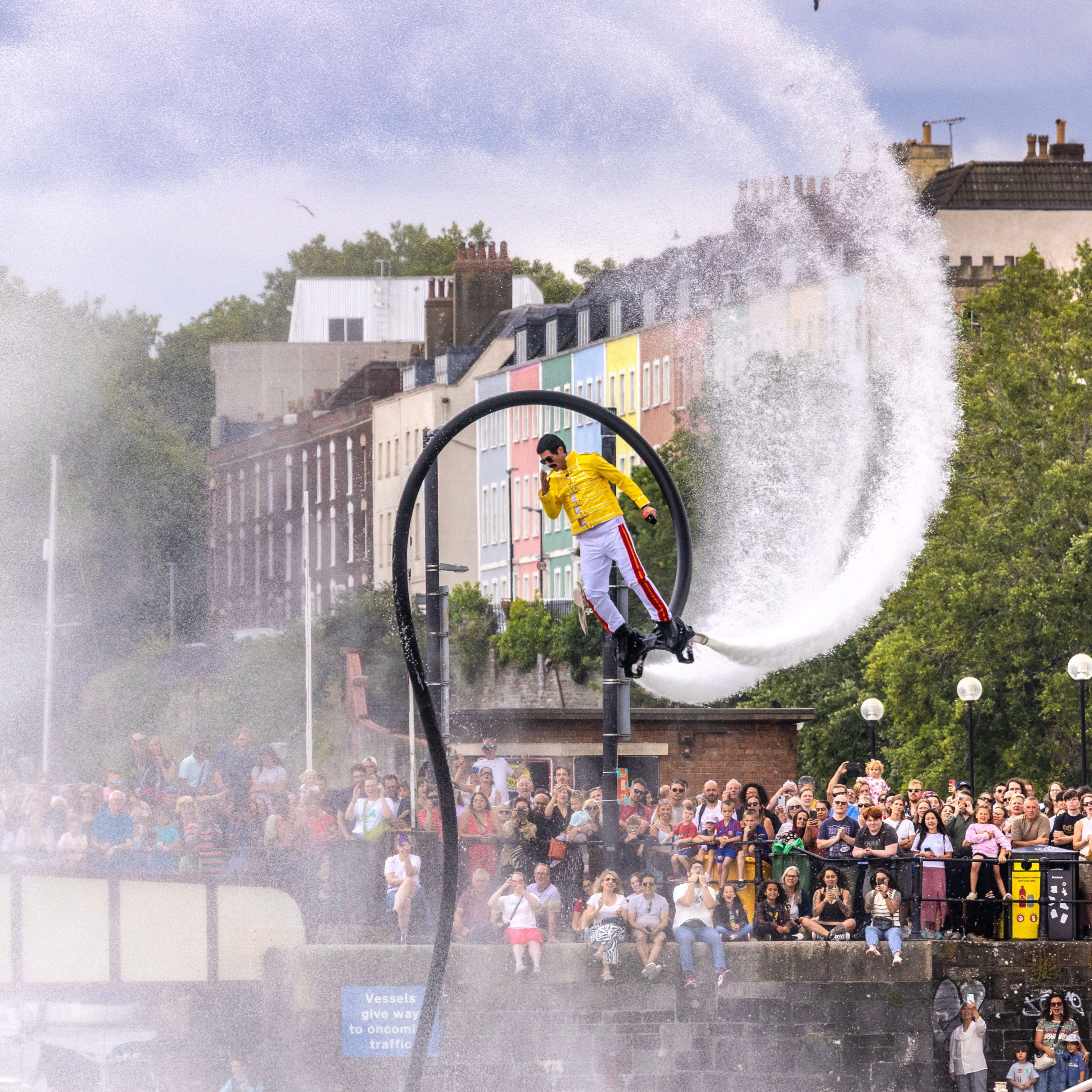 A performer dressed like Freddie Mercury flies above the water on a flyboard, spraying a high arc of water as a large crowd watches from the riverside.