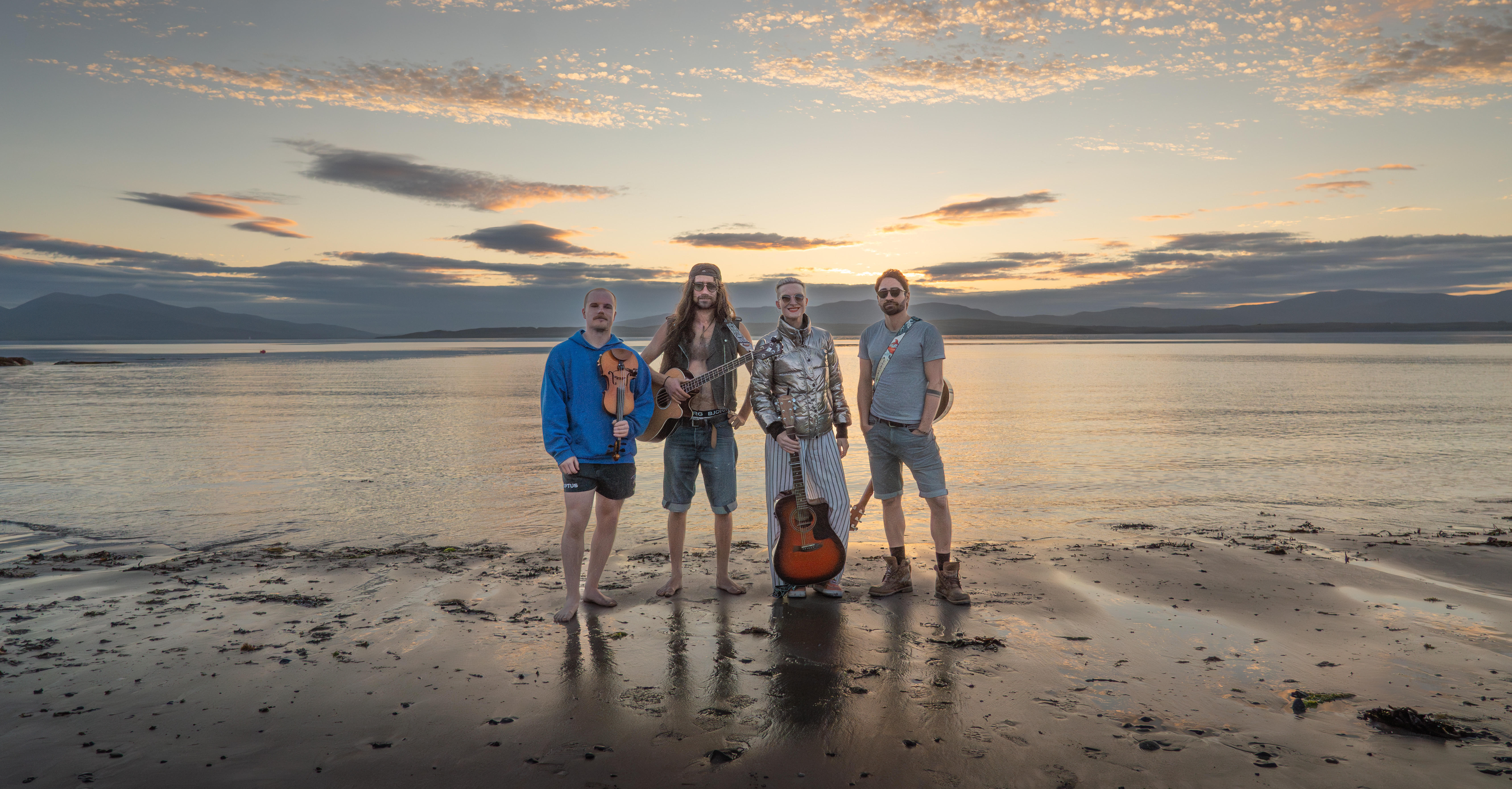 Four musicians stand on a beach at sunset holding instruments, with calm water and mountains in the background.