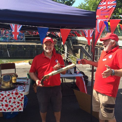 Two people in red shirts stand under a tent decorated with Union Jack bunting, one holding a historic-looking object, at an outdoor stall by the water.