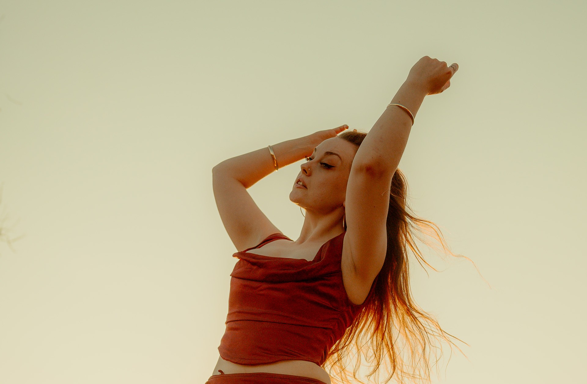 A person in a flowing red outfit poses with their arms raised against a glowing sunset sky, their hair catching the light in the breeze.