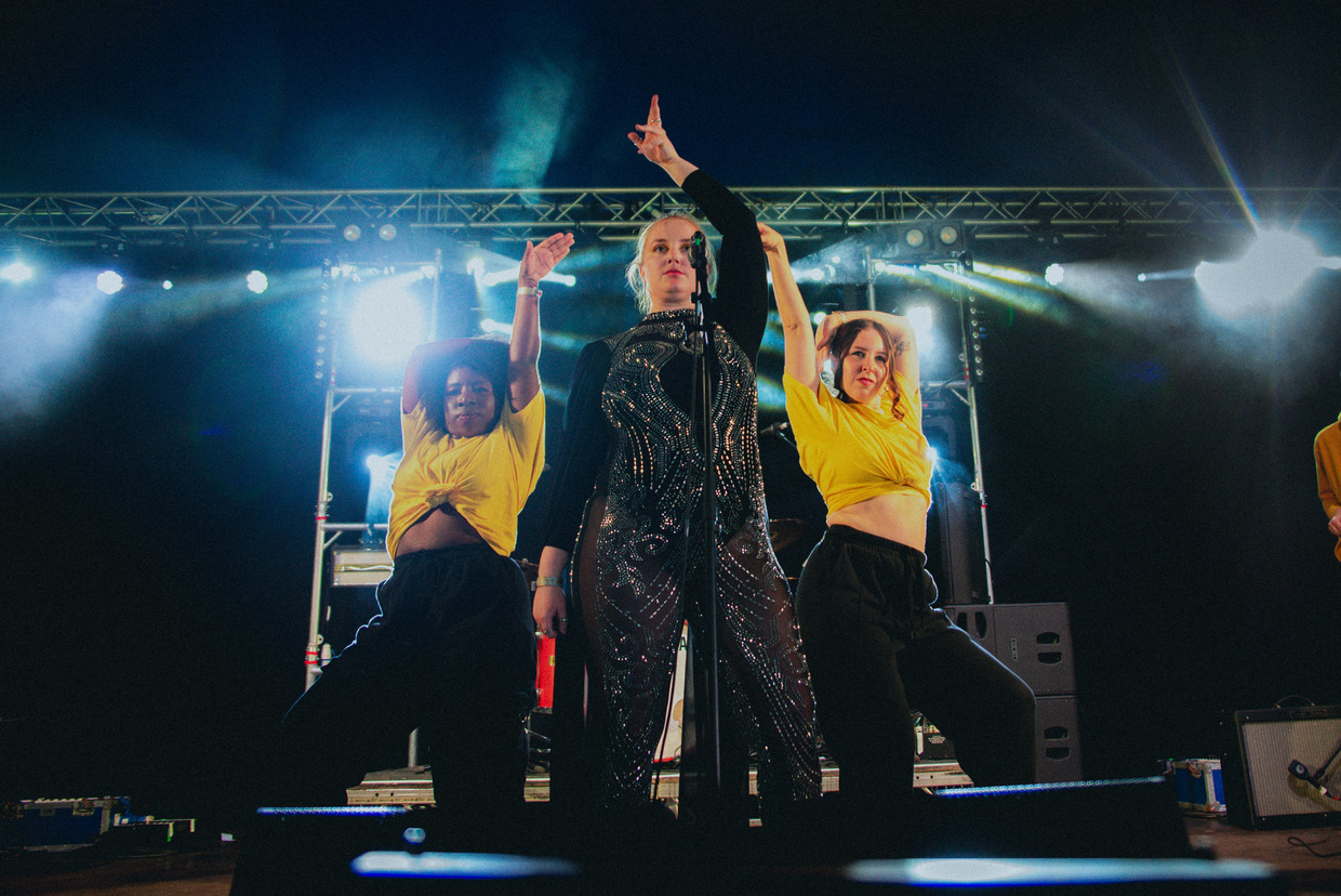 A singer in a black sequined outfit performs on stage between two dancers in yellow tops, posing under bright concert lights during a live nighttime performance.