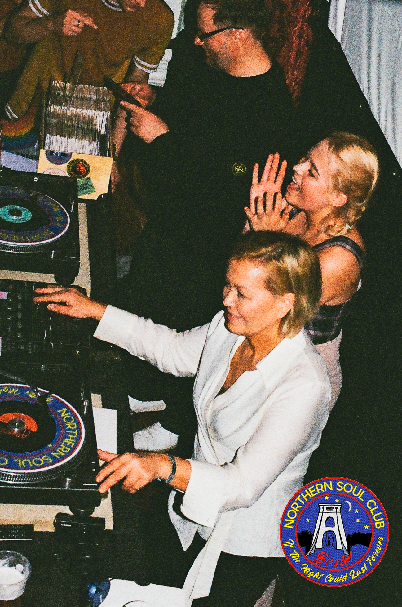 People dancing and DJing at a Northern Soul Club night in Bristol, with vinyl turntables, record boxes and partygoers gathered around the decks in a lively club atmosphere.