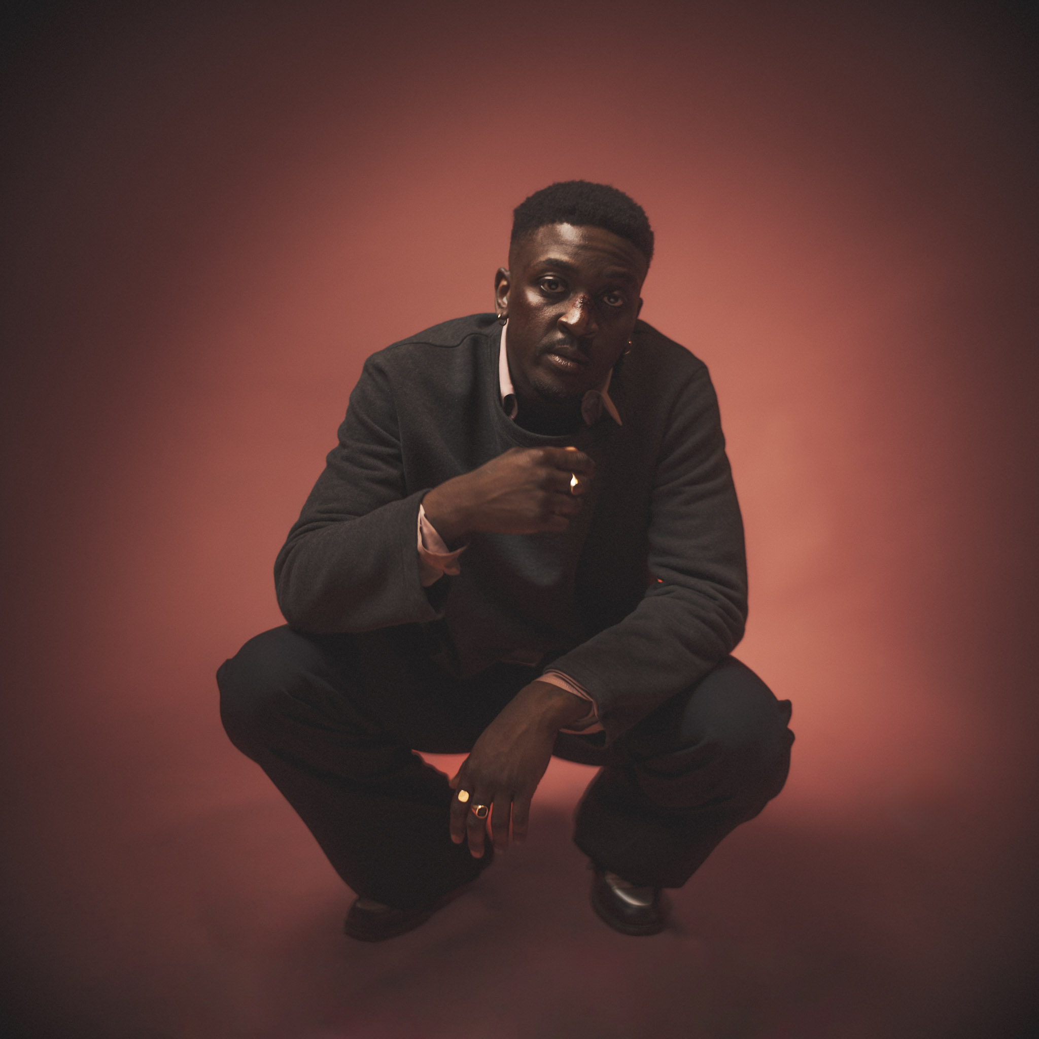 A man crouches against a warm pink and orange studio backdrop, wearing dark clothing and looking directly at the camera in a moody portrait photograph.