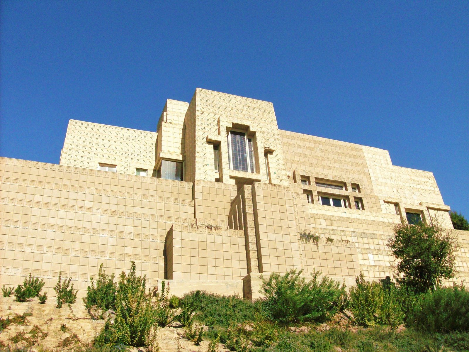 The Mayan-style concrete block walls of Ennis House rising above its hillside garden against a blue sky