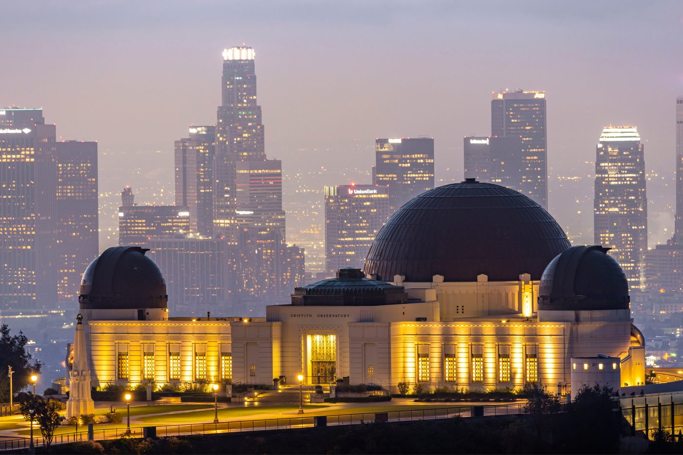 Griffith Observatory lit up at dusk, with the downtown Los Angeles skyline behind it