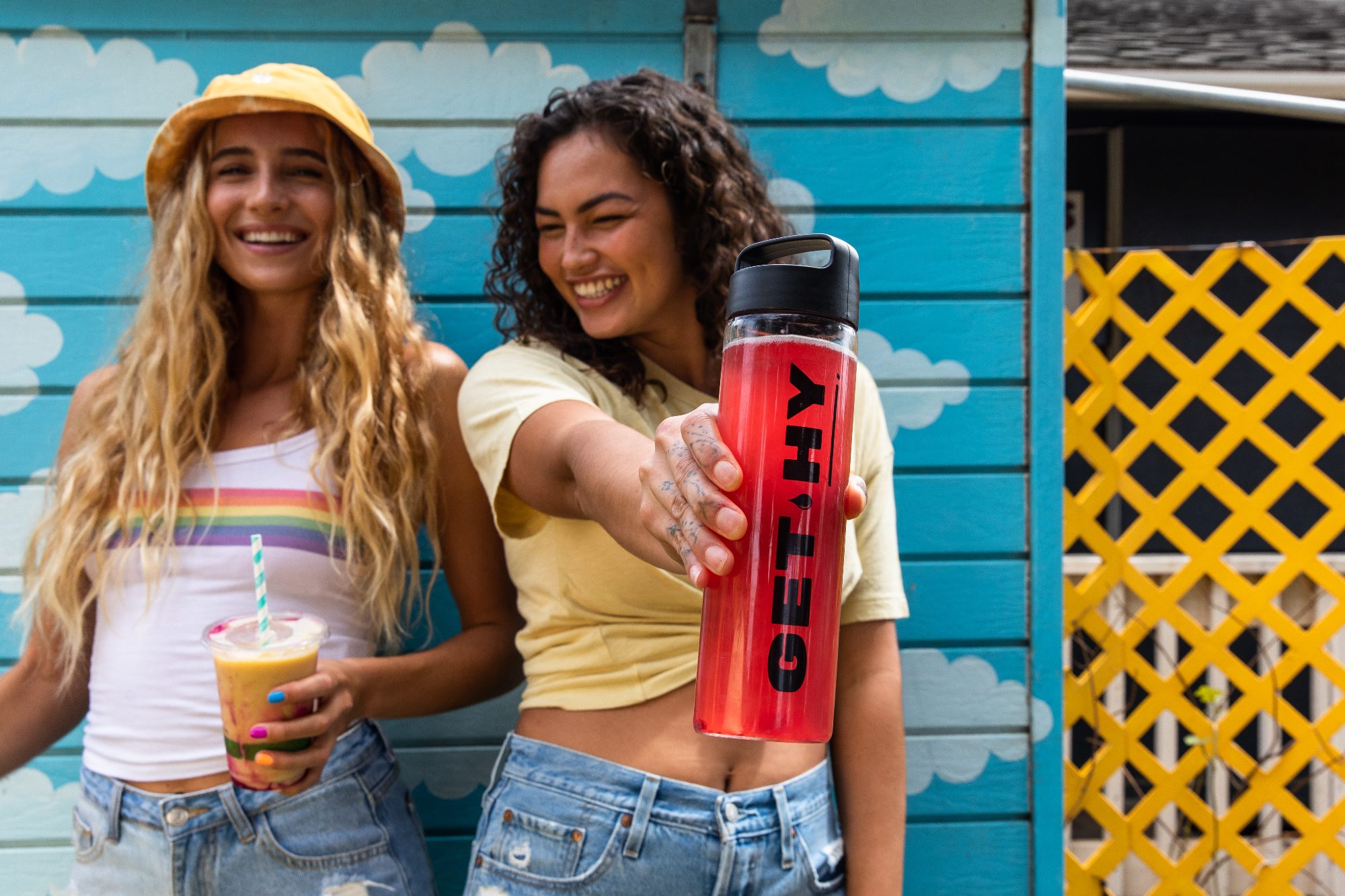 Two friends in front of a blue wall, one holding out a bottle
