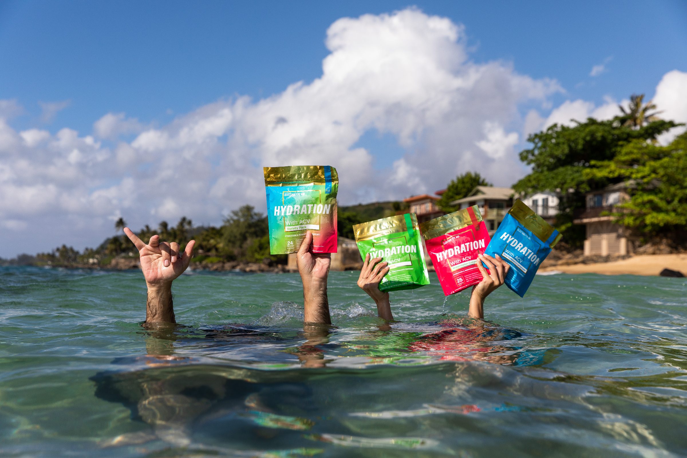 Four hands hold Essential Elements hydration packs up out of the sea in Hawaii