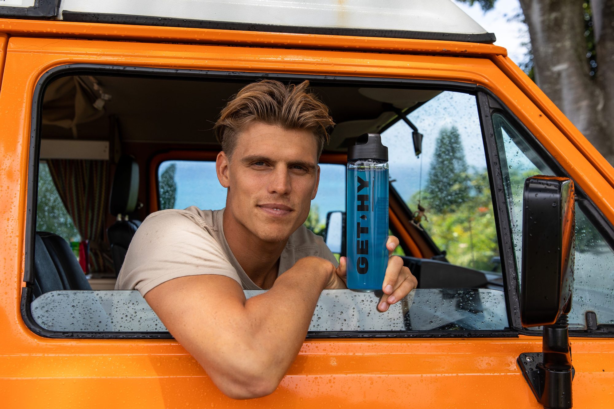 Koa Smith leans out of an orange van window holding an Essential Elements bottle
