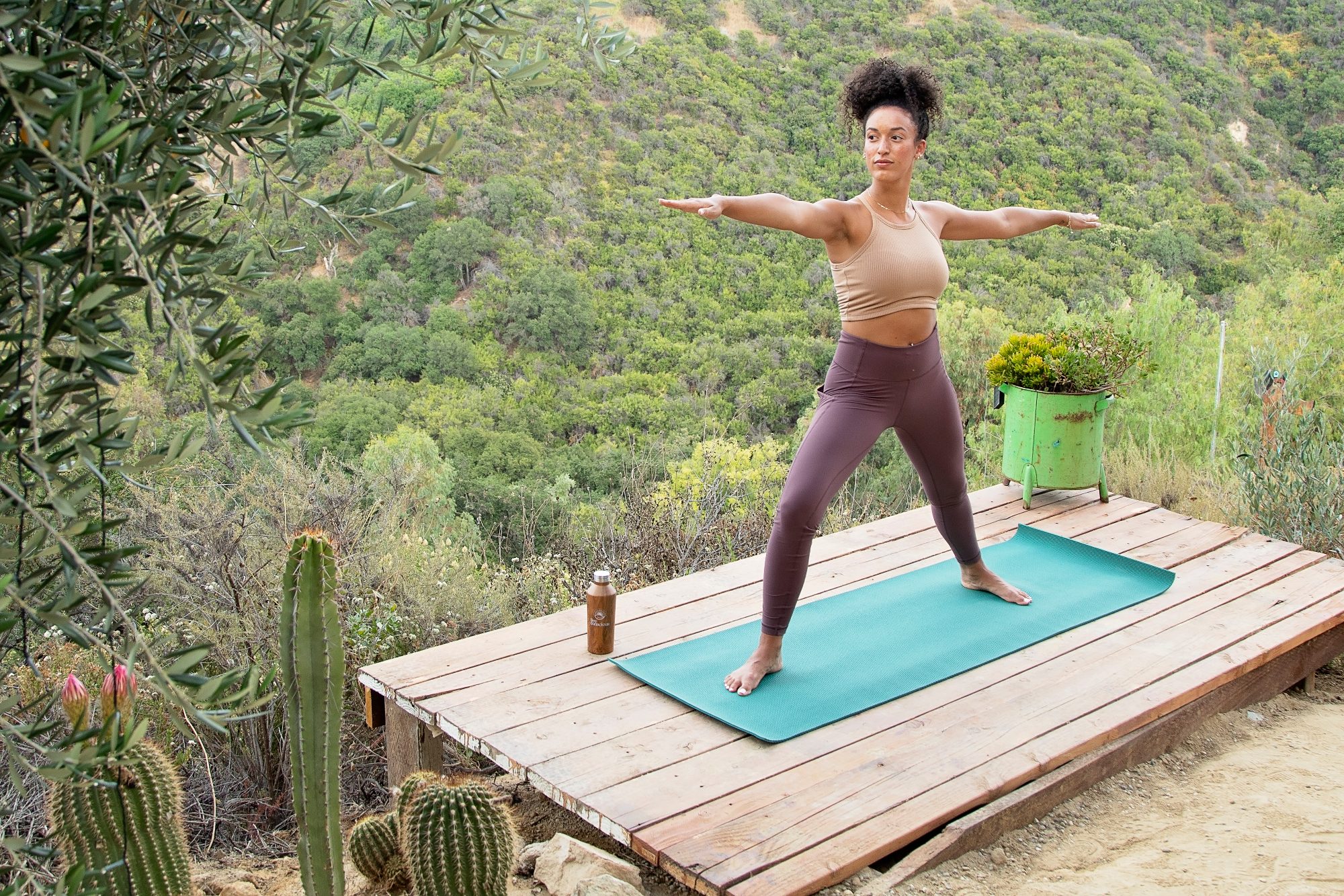 A woman holds a yoga pose on a wooden deck above a green valley