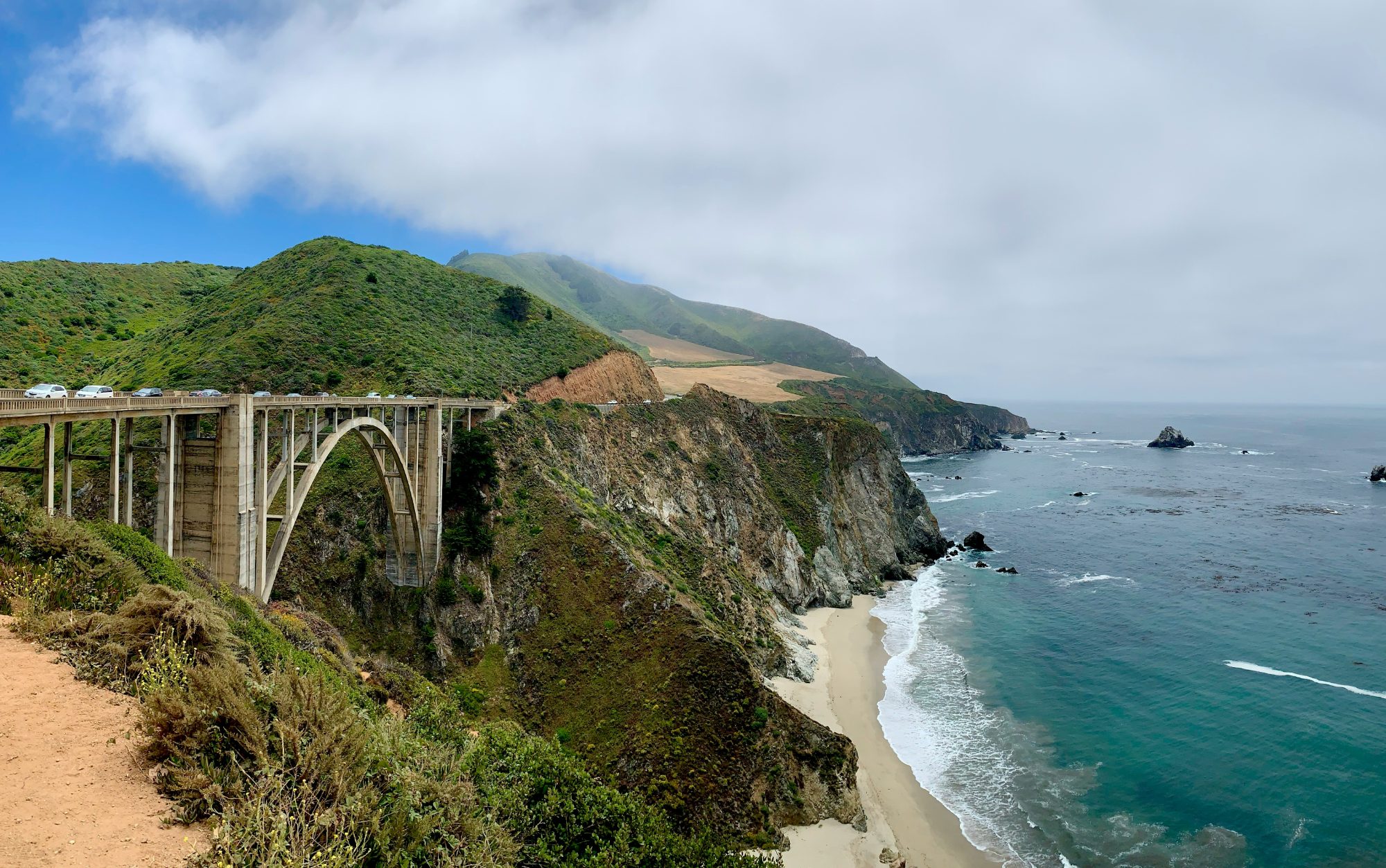 Bixby Creek Bridge, a tall concrete arch crossing a Big Sur canyon, with green coastal hills and a turquoise ocean beyond