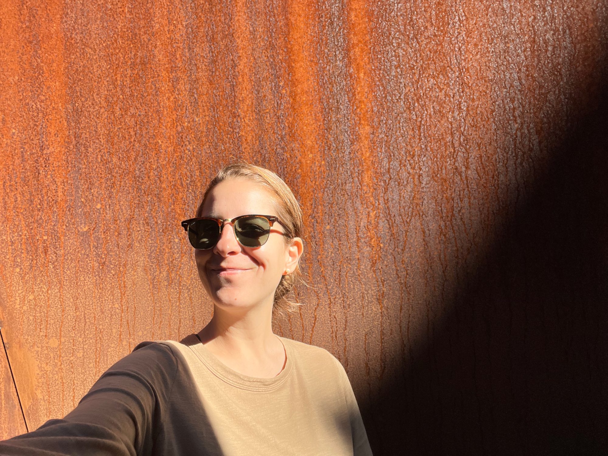 Valentina smiling in sunglasses inside Richard Serra's Sequence, with a tall wall of rust-brown weathering steel behind her