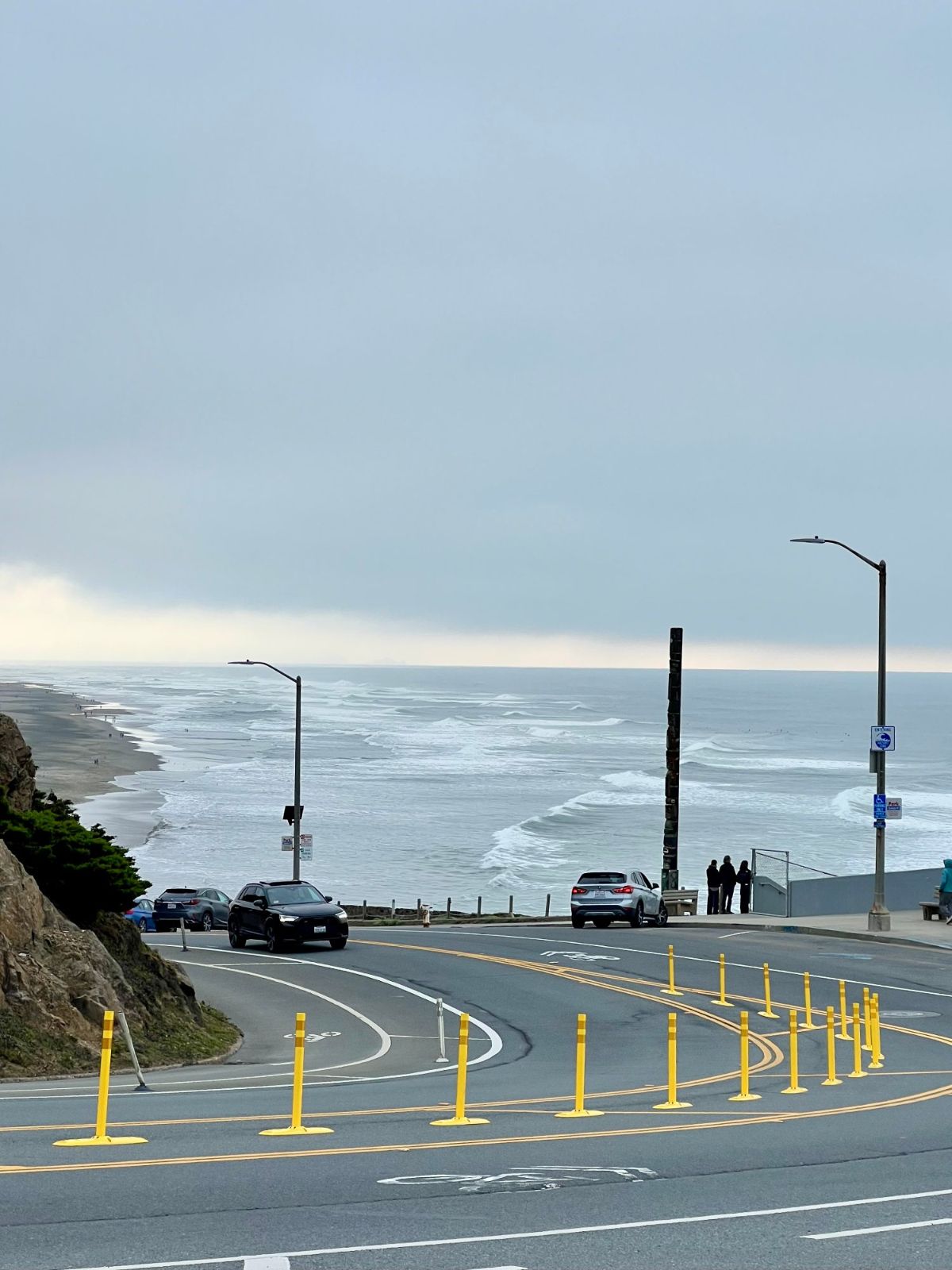 A curving road above Ocean Beach lined with yellow posts, with waves breaking in the distance