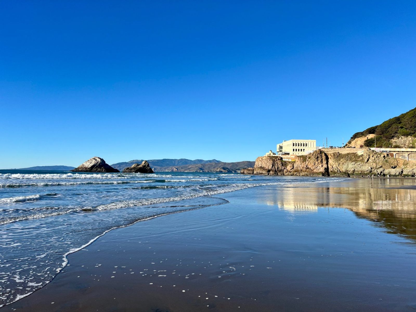 A wet, mirror-like beach at the north end of Ocean Beach, with the Cliff House on the headland at the right