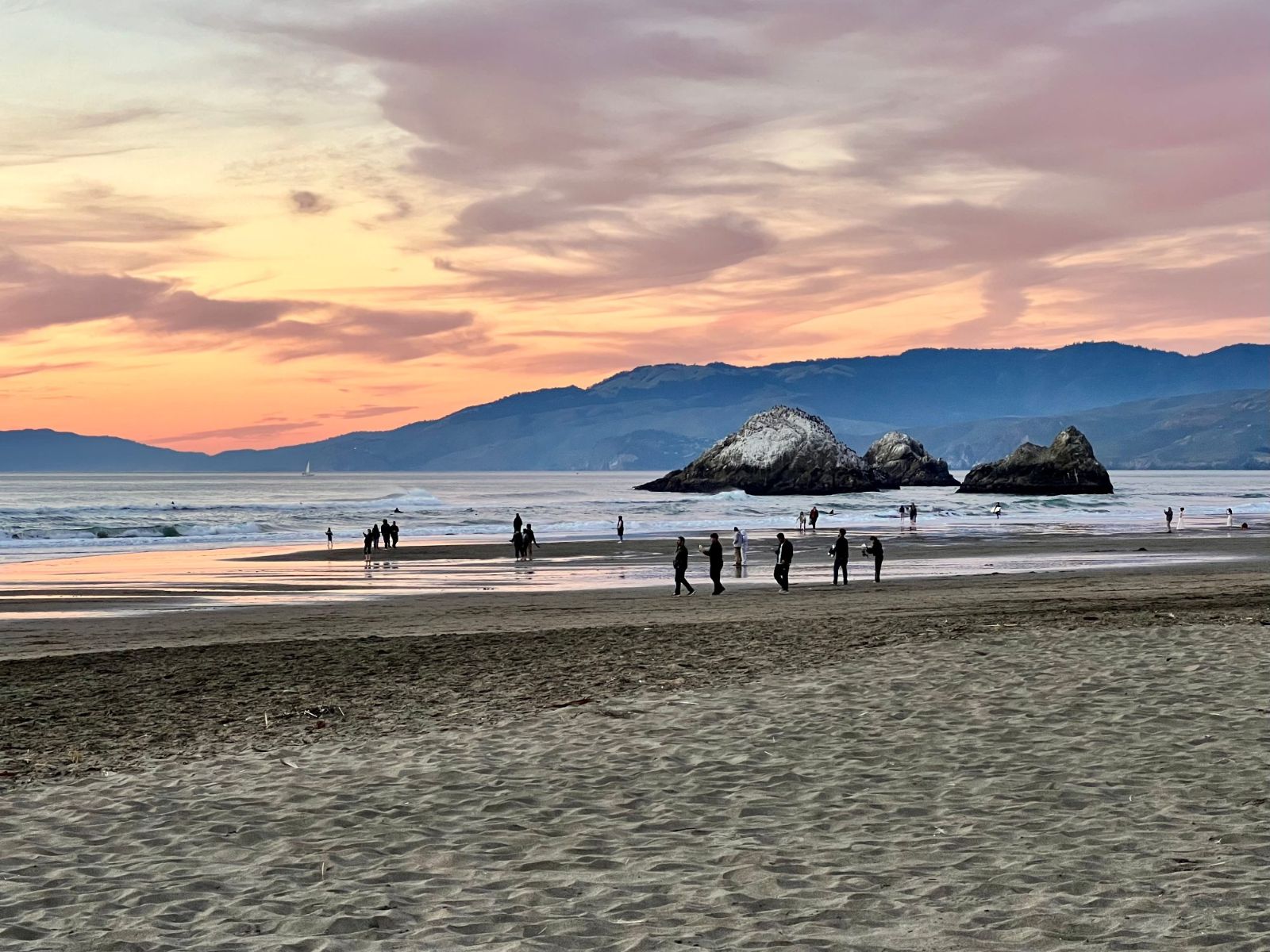 Golden-hour light on Ocean Beach, with people at the waterline and an offshore rock under a pink sky