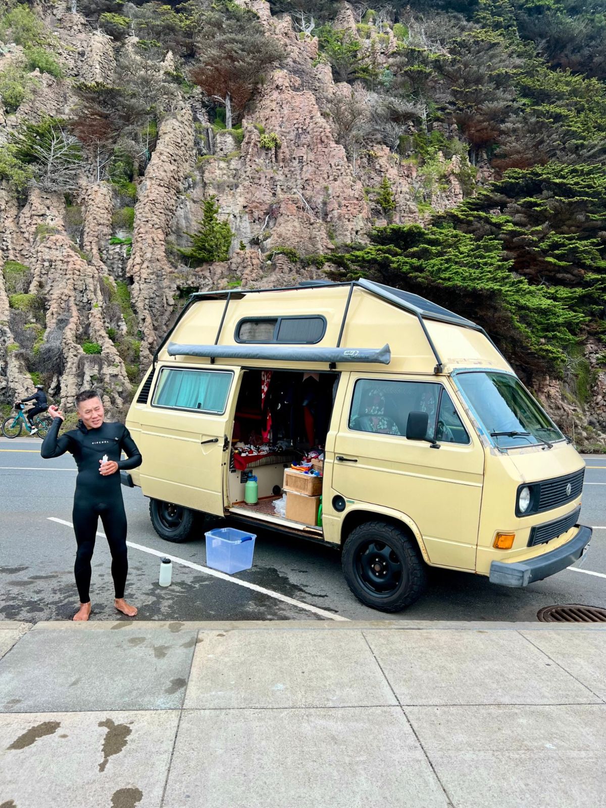 A surfer in a wetsuit smiling beside a cream-colored camper van parked below a green hillside