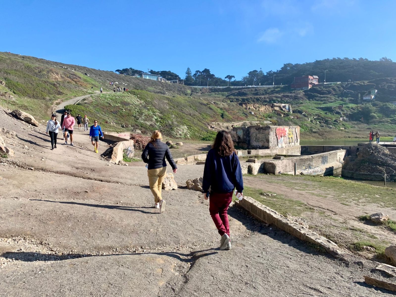 Visitors walking down a dirt and concrete path toward the Sutro Baths ruins under a blue sky