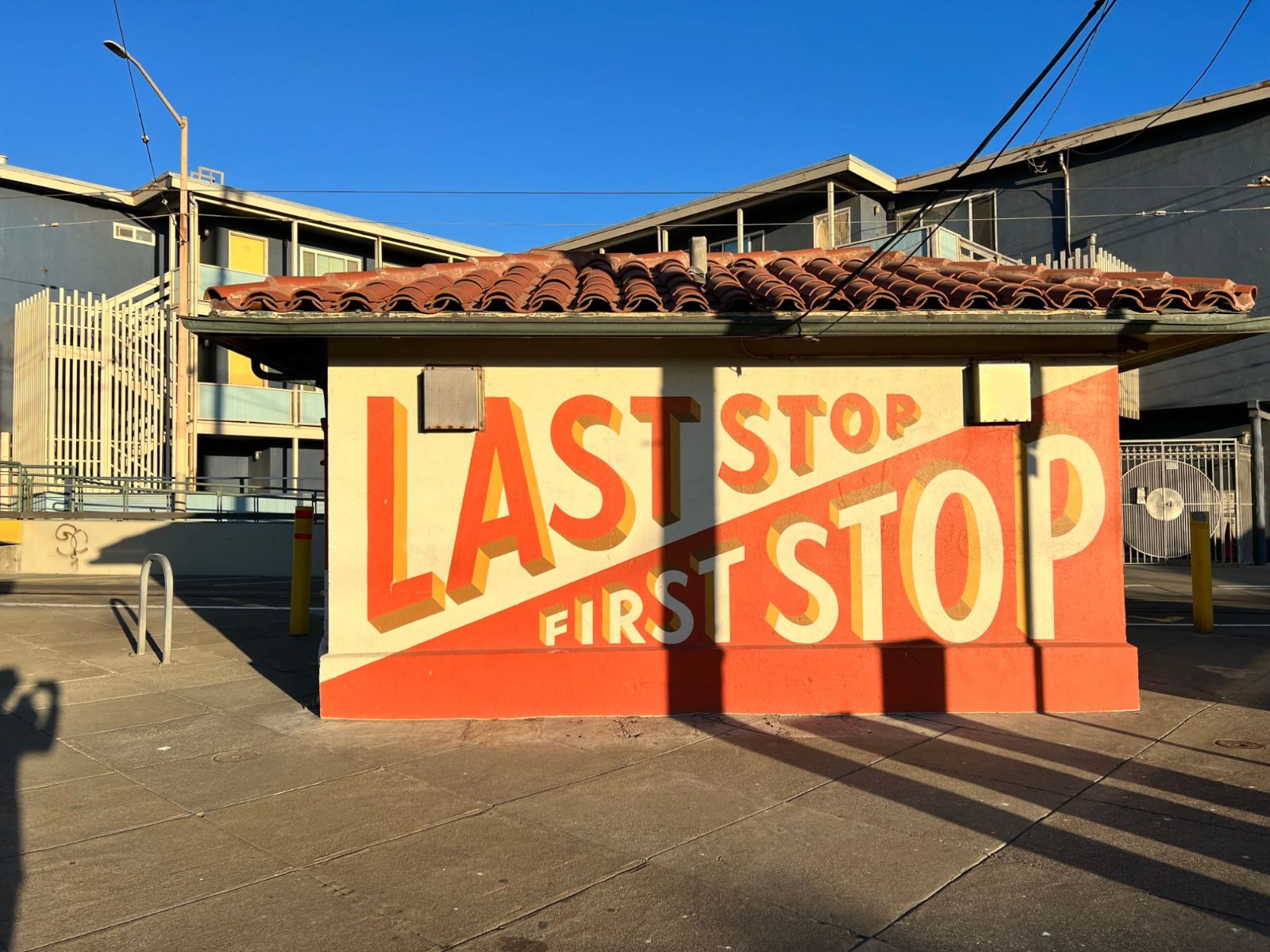 A hand-lettered red and cream kiosk reading Last Stop and First Stop, with apartment buildings behind it