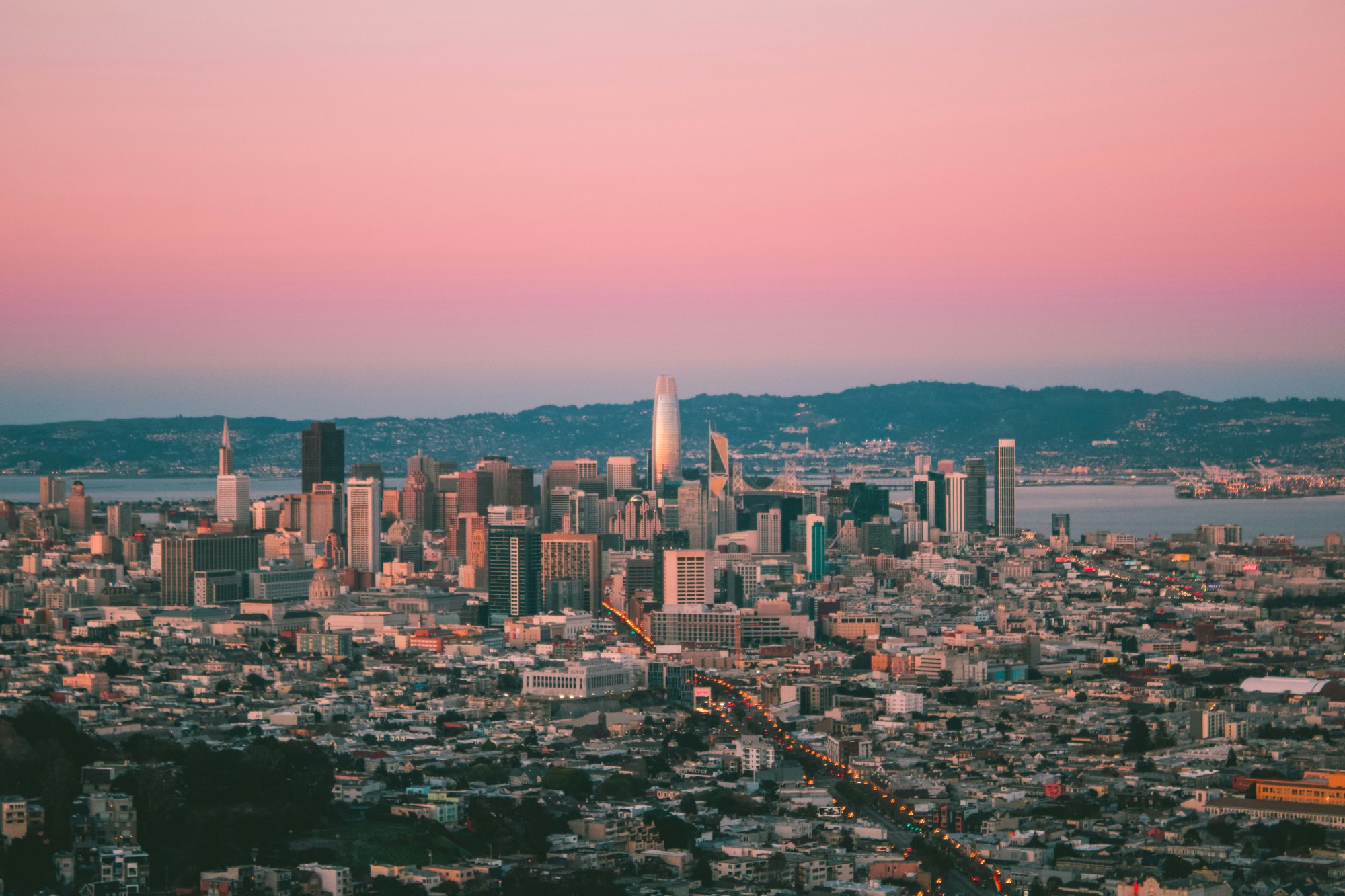 San Francisco's downtown skyline at dusk under a pink sky, with the Salesforce Tower catching the last sun and the Bay Bridge beyond