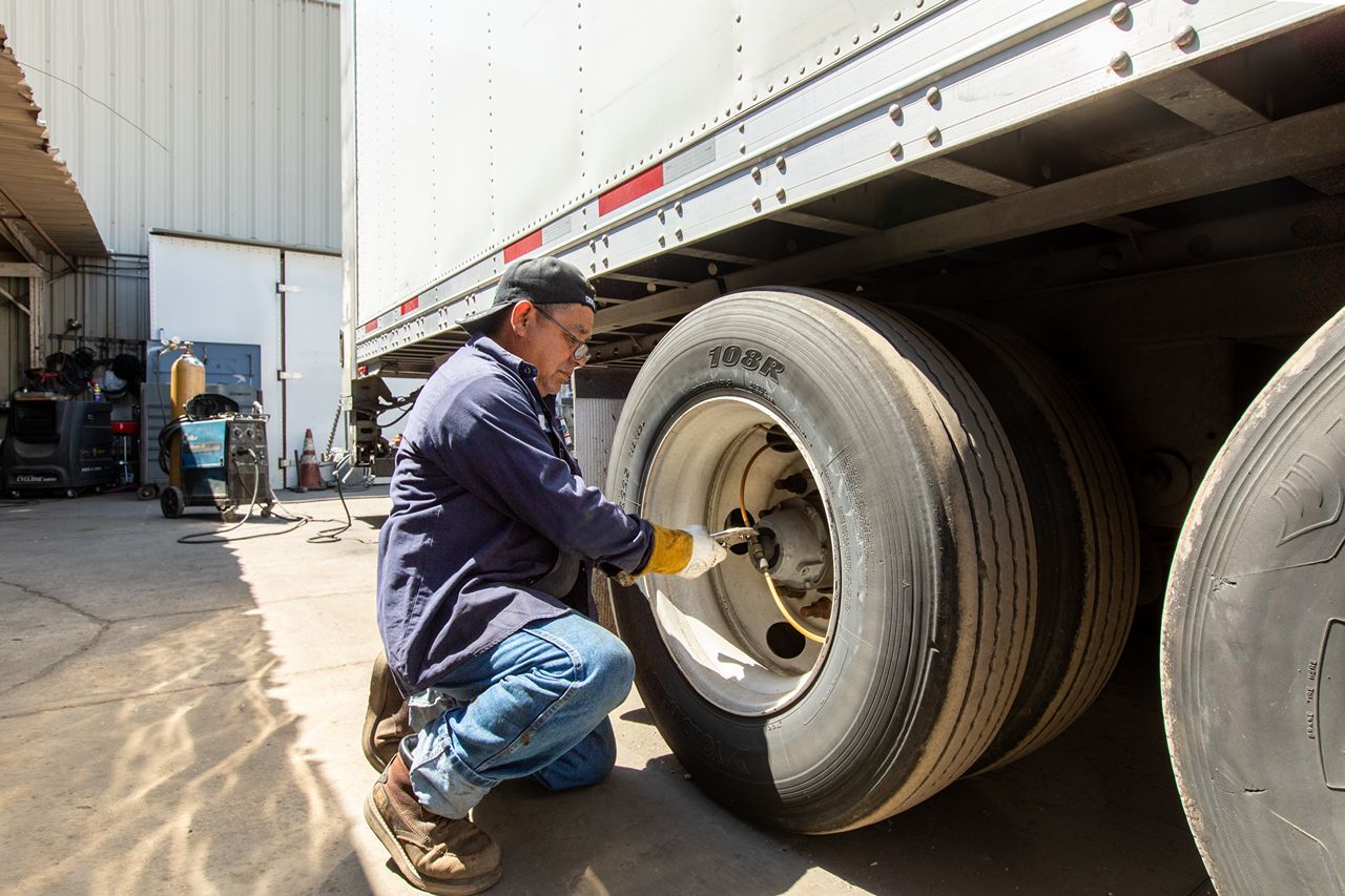Mechanic wearing gloves and cap repairs a trailer wheel with a power tool outside a truck service shop on sunny day.