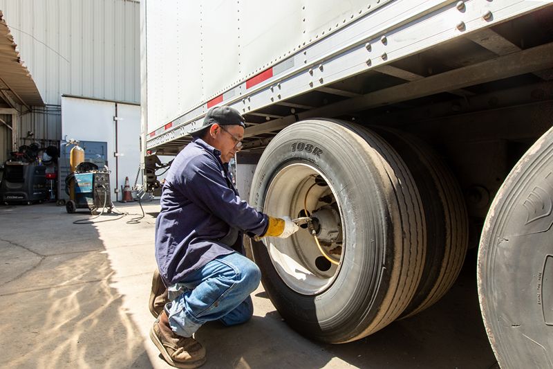 Mechanic wearing gloves and cap repairs a trailer wheel with a power tool outside a truck service shop on sunny day.