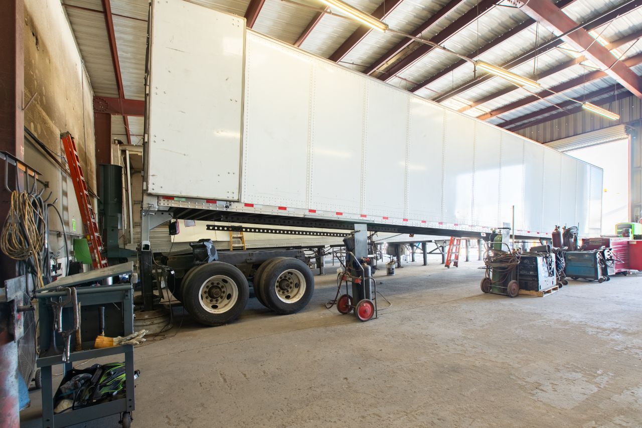 Large semi-trailer inside a repair shop, elevated for maintenance, surrounded by tools, welding equipment, and ladders under bright overhead lighting.